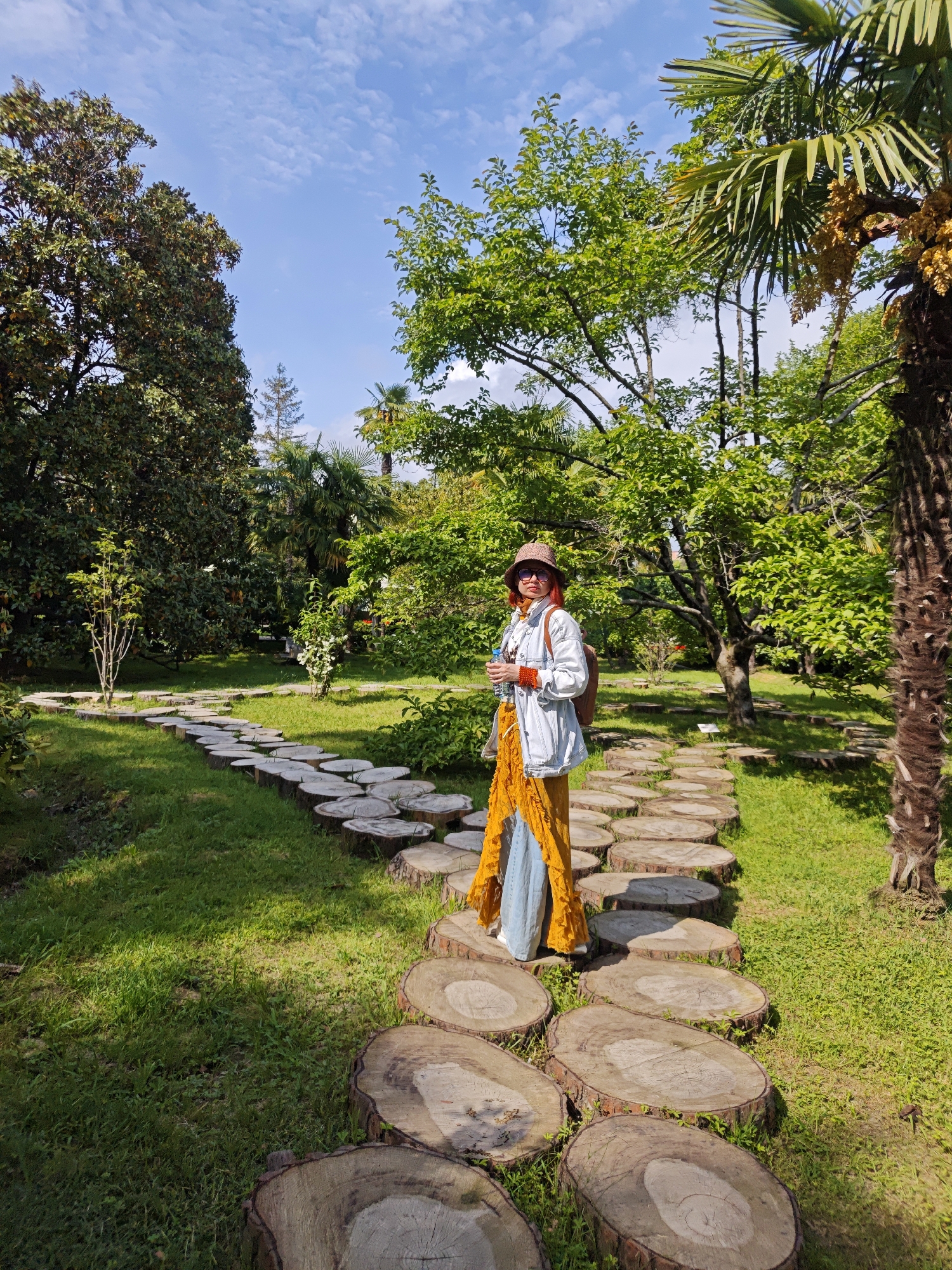 A full shot captures a woman standing on a path made of tree trunk slices in Sochi, Russia. She faces the viewer, wearing a hat, sunglasses, and a light denim jacket. A long, layered dress in yellow and light blue is visible. She also has a backpack. The pathway stretches through a grassy area dotted with trees and shrubs, leading into the distance. Palm trees and other greenery surround the area under a partially cloudy sky.