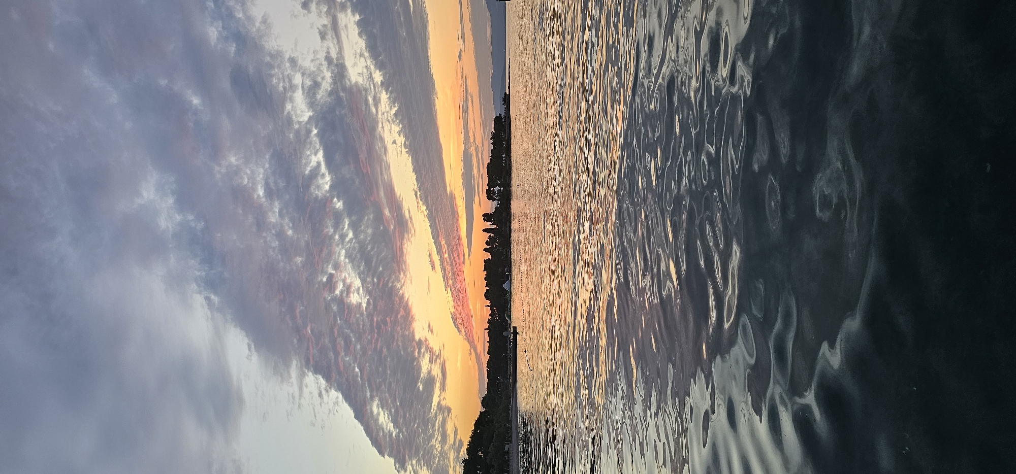 The image shows a sunset over the sea in Supetar, Croatia. The sky is filled with dramatic clouds, painted in hues of orange, pink, and purple, reflecting on the rippling water. In the distance, a silhouette of trees and buildings lines the coast, suggesting a settlement. The foreground is dominated by the dark, textured surface of the water, disturbed by gentle waves that catch the last light of the day. A few small buoys or markers are visible on the water's surface. The overall scene is serene and evokes the end of a day.