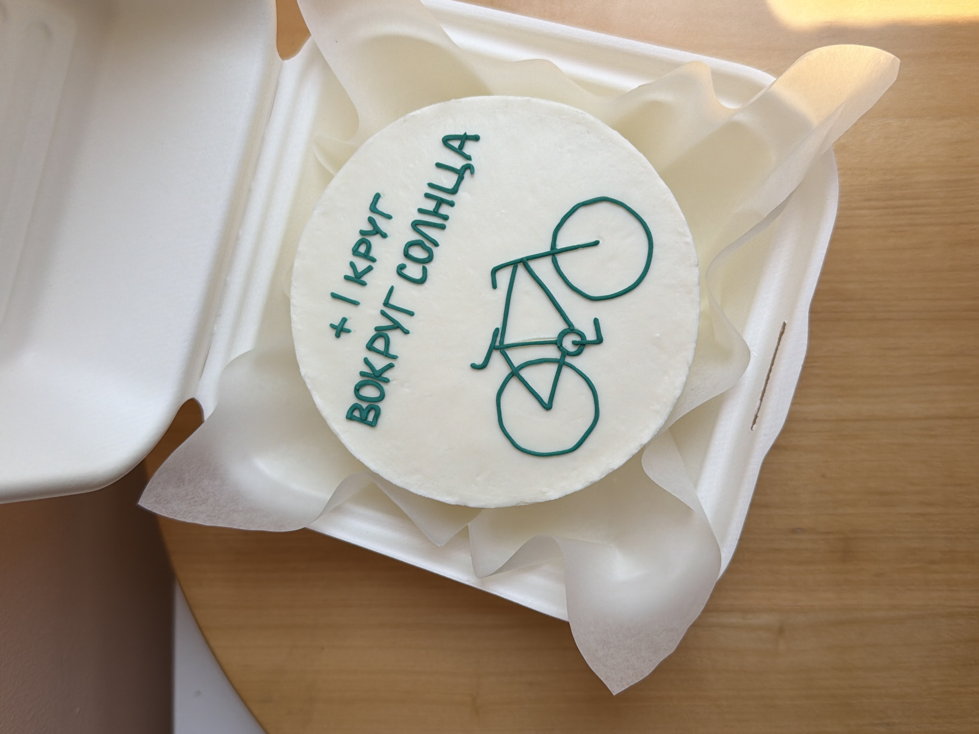 A close-up, top-down shot depicts a round white cake decorated with a green drawing of a bicycle and green Cyrillic text, nestled in a white takeout container lined with white tissue paper. The container sits on a light brown wooden surface. The cake appears to be a celebration cake. The Cyrillic text translates to "+ 1 circle around the sun." The cake may have been purchased from a bakery in Israel.