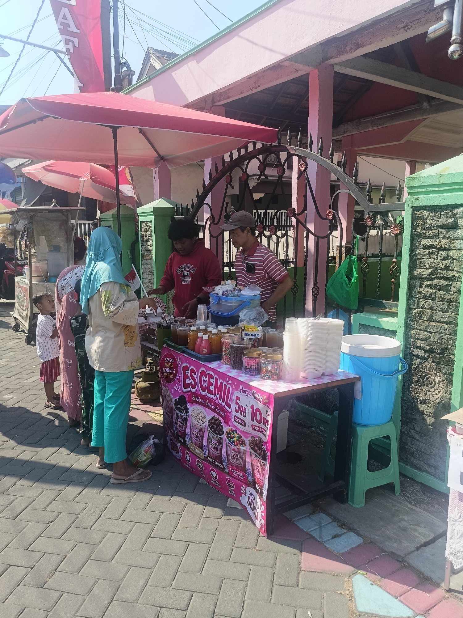 In the city of Gedangan, Indonesia, a street vendor is serving customers at a portable stall. The stall is set up on a paved sidewalk under a red umbrella. A pink banner on the front of the stall advertises "ES CEMIL" with images of ice cream and prices. Behind the stall, a wrought iron gate separates the sidewalk from a building with a pink exterior and stone accents.
Two men are working behind the stall. One, wearing a red hoodie with "DSENZA 1997" printed on it, is handing something to a customer. The other man, wearing a striped polo shirt and a cap, is partially visible to the right.
Three customers are at the stall. A woman wearing a light-colored headscarf and teal pants is facing the vendor, holding a small white flag. A child in a striped shirt and red shorts stands to her left, looking up at the vendor. Another woman in a patterned pink outfit is partially obscured behind the woman in teal.
On the stall, there are several jars of colorful sprinkles, bottles of liquid, stacks of white plastic cups, and a large blue cooler. A green plastic chair is visible to the right of the cooler. The environment is bright and sunny, with power lines visible overhead.