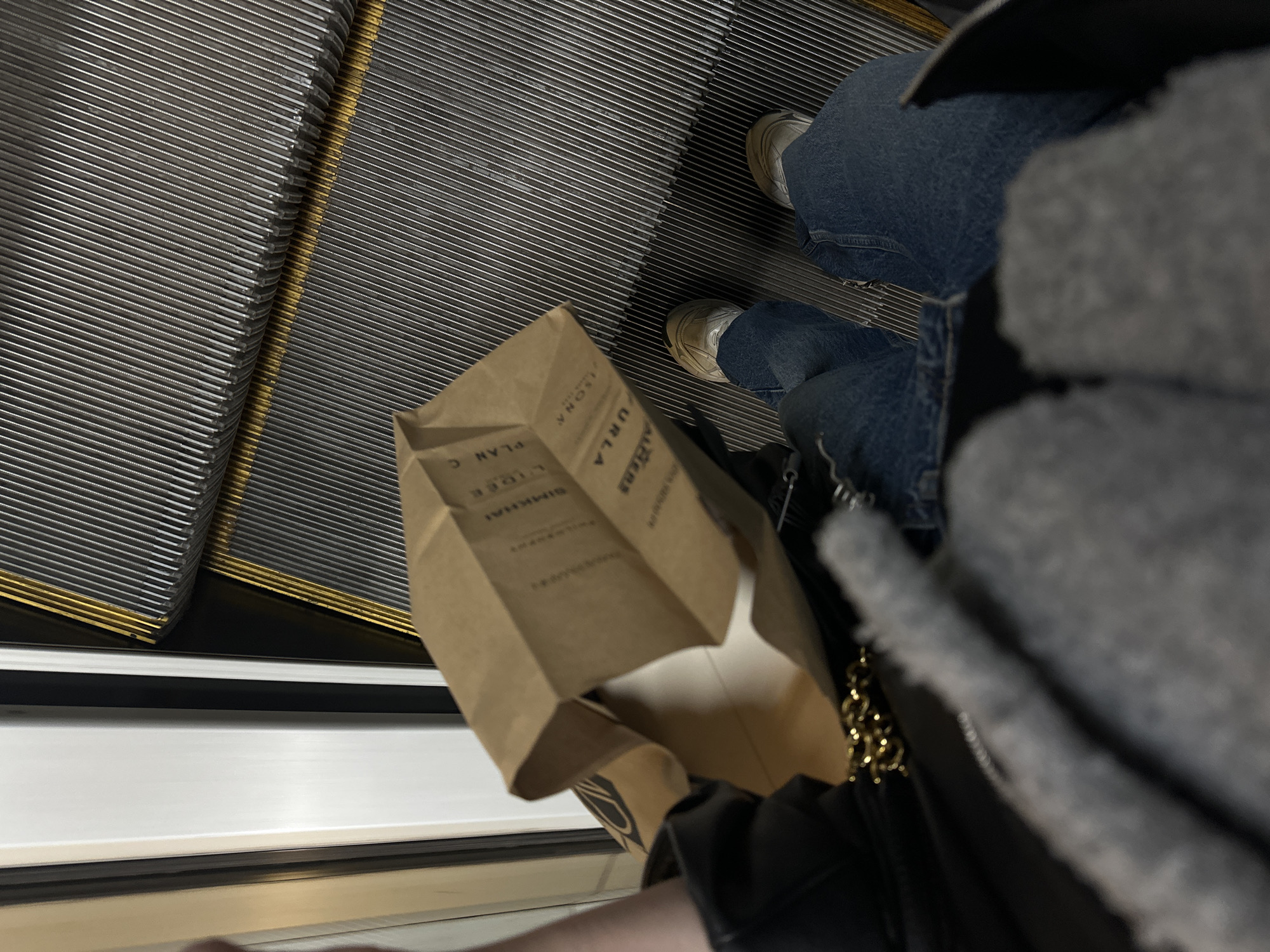 The image captures a first-person perspective looking down at a person riding an escalator in what appears to be an indoor public setting, likely a shopping mall or a metro station in Sofia, Bulgaria. The main subject, partially visible from the mid-thigh down, is wearing blue jeans and light-colored sneakers with thick soles. A dark jacket or coat with a grey, possibly faux fur or shearling, lining is visible on the right, along with a gold chain strap from a bag.
In the foreground, a brown paper bag rests near the person's feet. The bag features multiple brand names and words printed vertically in black text. Clearly visible are "FURLA," "PLAN C," "LIGÉE," and "SIMKHAI." Other partially visible words include "ALBOS" (or "ALBDS"), "SONNA" (or "ISONNA"), "ONA," and "RUD," with "PAMPANINI" faintly discernible below "SIMKHAI."
The escalator steps are made of ridged, metallic gray material. A distinct yellow safety stripe runs along the edge of each step, providing a visual guide. The side panel of the escalator is smooth, metallic, and light-colored, reflecting the ambient artificial light of the indoor environment. The image is taken from a high angle, emphasizing the pattern of the escalator steps and the items carried by the individual. No specific time of day or weather conditions are discernible, consistent with an indoor setting.