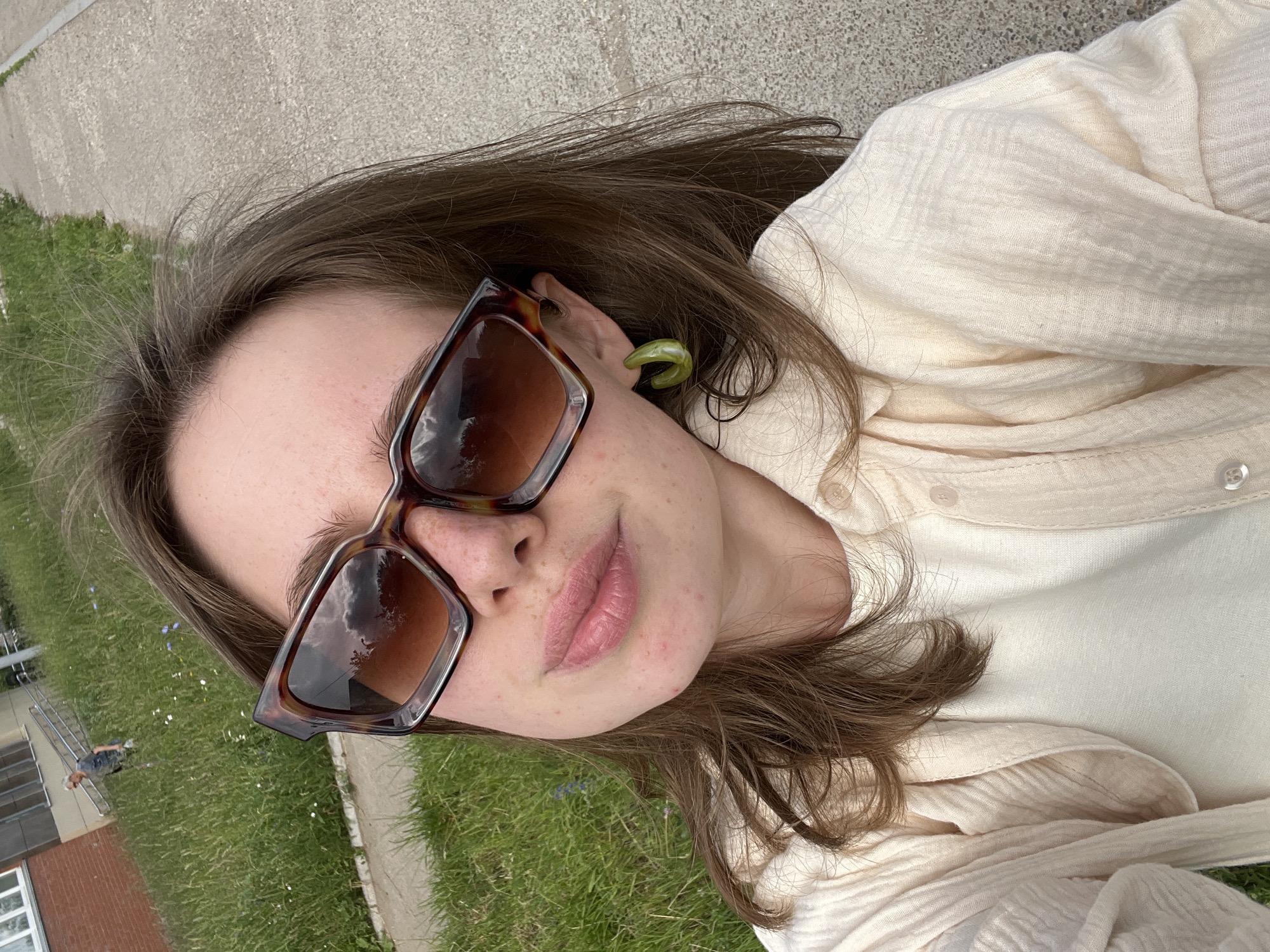 A young woman with long brown hair, wearing large tortoiseshell sunglasses, a light-colored collared shirt over a white t-shirt, and green hoop earrings, poses for a selfie outdoors. She is positioned in the foreground, with a paved pathway and a grassy area with scattered wildflowers behind her. In the background, a brick building with a staircase leading to an entrance is visible, and a person is walking on the grass. The overall scene suggests a casual outdoor setting in Chaikovsky, Russia.