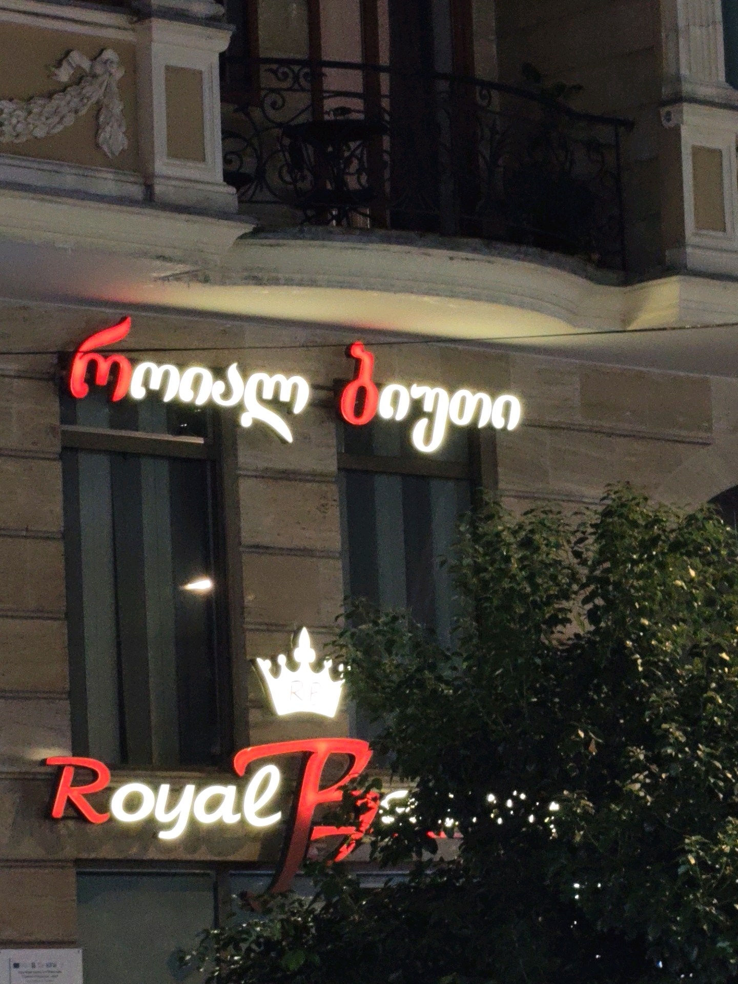 The image shows a nighttime exterior of a building in Batumi, Georgia. A neon sign in the Georgian alphabet, glowing red and white, is prominently displayed. The text appears to read "რკონის ბაღი" (Rkonis Baghi), which translates to "Rkoni Garden". Below this, another neon sign in English reads "Royal P" in red and white letters, with a white crown emblem above it. The building has architectural details, including ornate molding and balconies. Lush green trees are visible in the foreground, partially obscuring the lower portion of the signs. The lighting suggests it is after dark, and the scene is likely a commercial establishment.