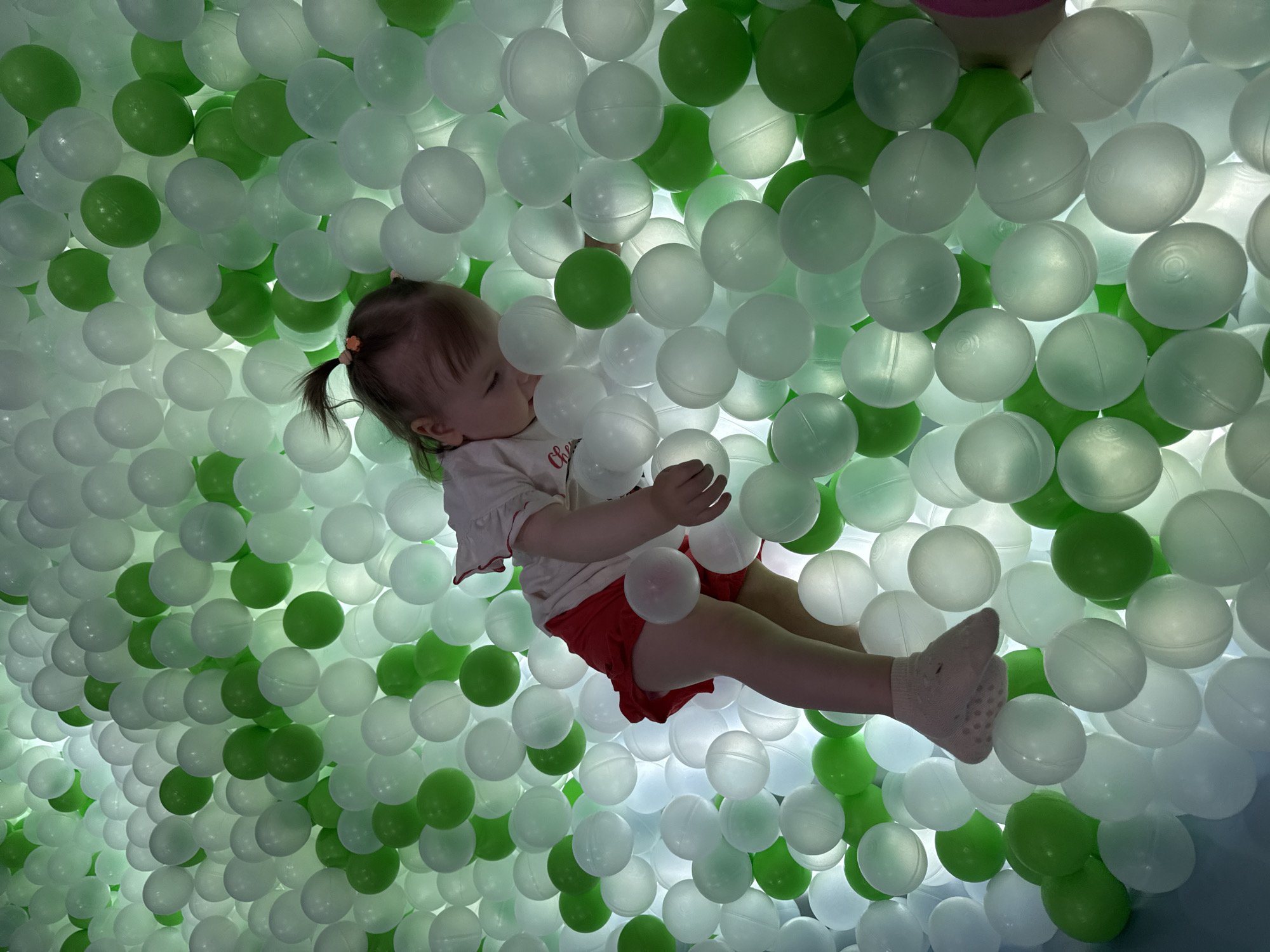 A young child sits in a ball pit filled with white and green plastic balls. The child, wearing a white shirt, red shorts, and socks, is seen from an overhead perspective. The environment is illuminated, with the light seemingly coming from beneath the balls. The exact location is not discernible from the image. The scene suggests a play area intended for children.