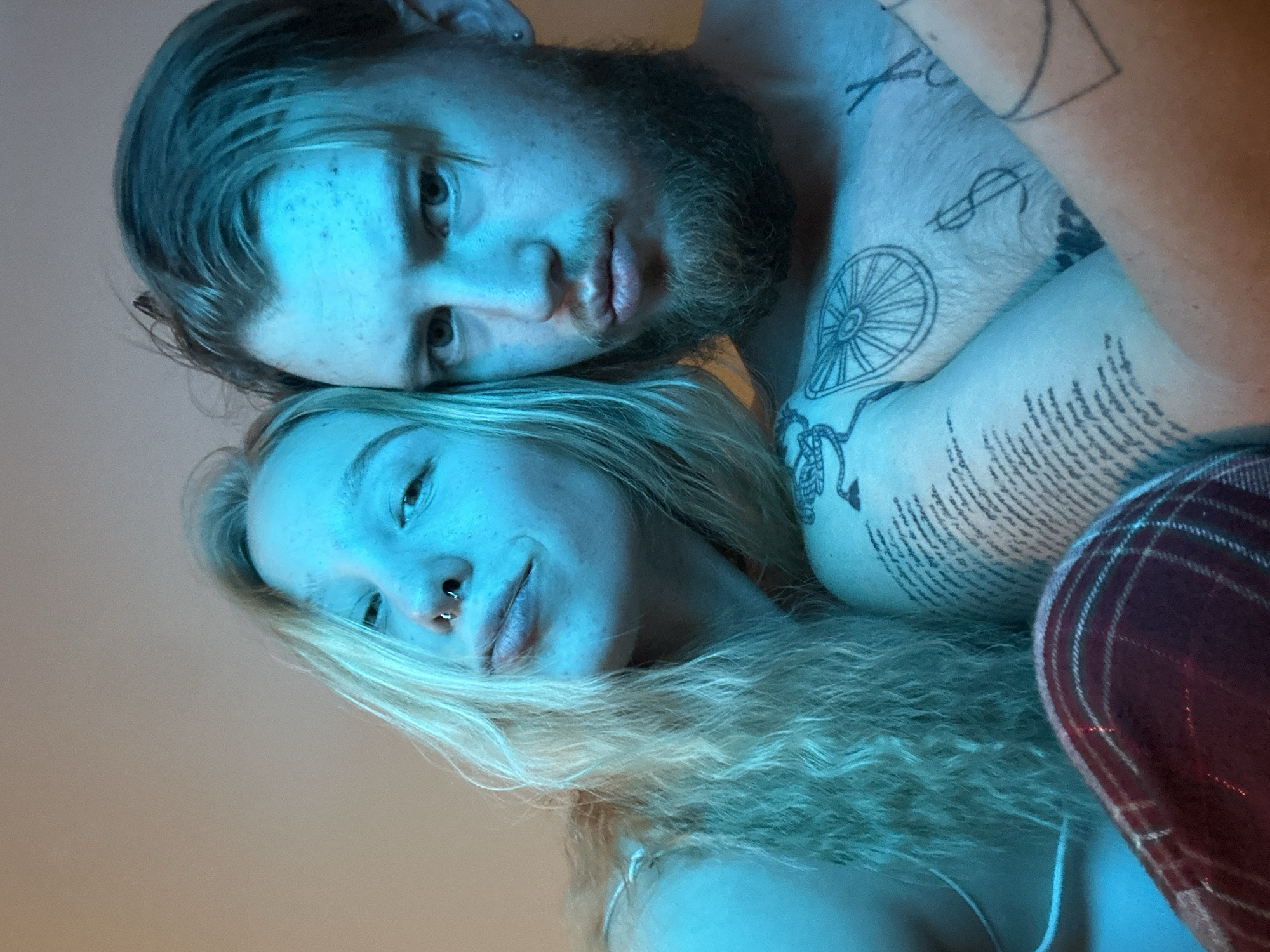 A close-up photograph depicts a fair-skinned man with a dark beard and a fair-skinned woman with blonde, wavy hair in an intimate pose. The man is positioned above the woman, his head near hers. The lighting is predominantly blue. The man has tattoos on his chest and arms, including a wheel-like design and text. The woman has a nose ring. Plaid fabric is visible in the lower right corner. The location is unknown and unidentifiable from the image. The city of Budva, Montenegro is not visually suggested.