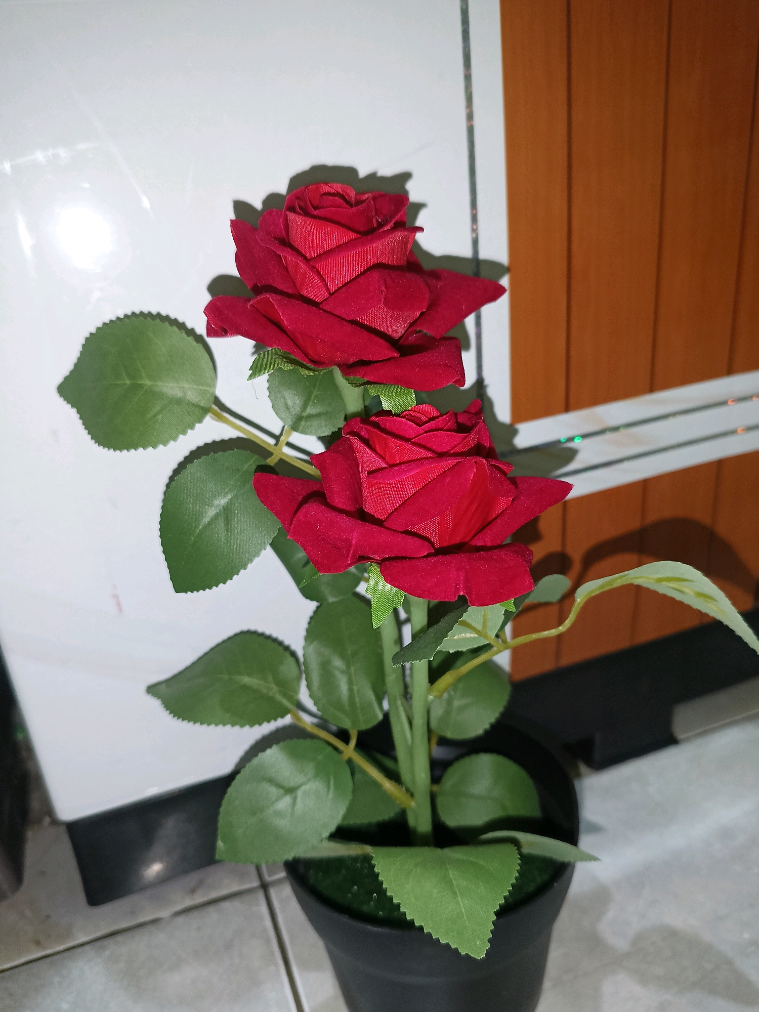 A close-up, eye-level shot features a faux potted red rose plant. The plant has two prominent red rose blooms, each with layered petals, and several green leaves with serrated edges. The plant is housed in a black pot, the top of which is visible. The background consists of a white tiled wall on the left, with a bright light source casting reflections, and a brown wooden paneled wall on the right. The floor appears to be tiled with light gray and white tiles. The scene is indoors, in an area that could be a living room or a decorative corner of a home or establishment in Tangerang, Indonesia.