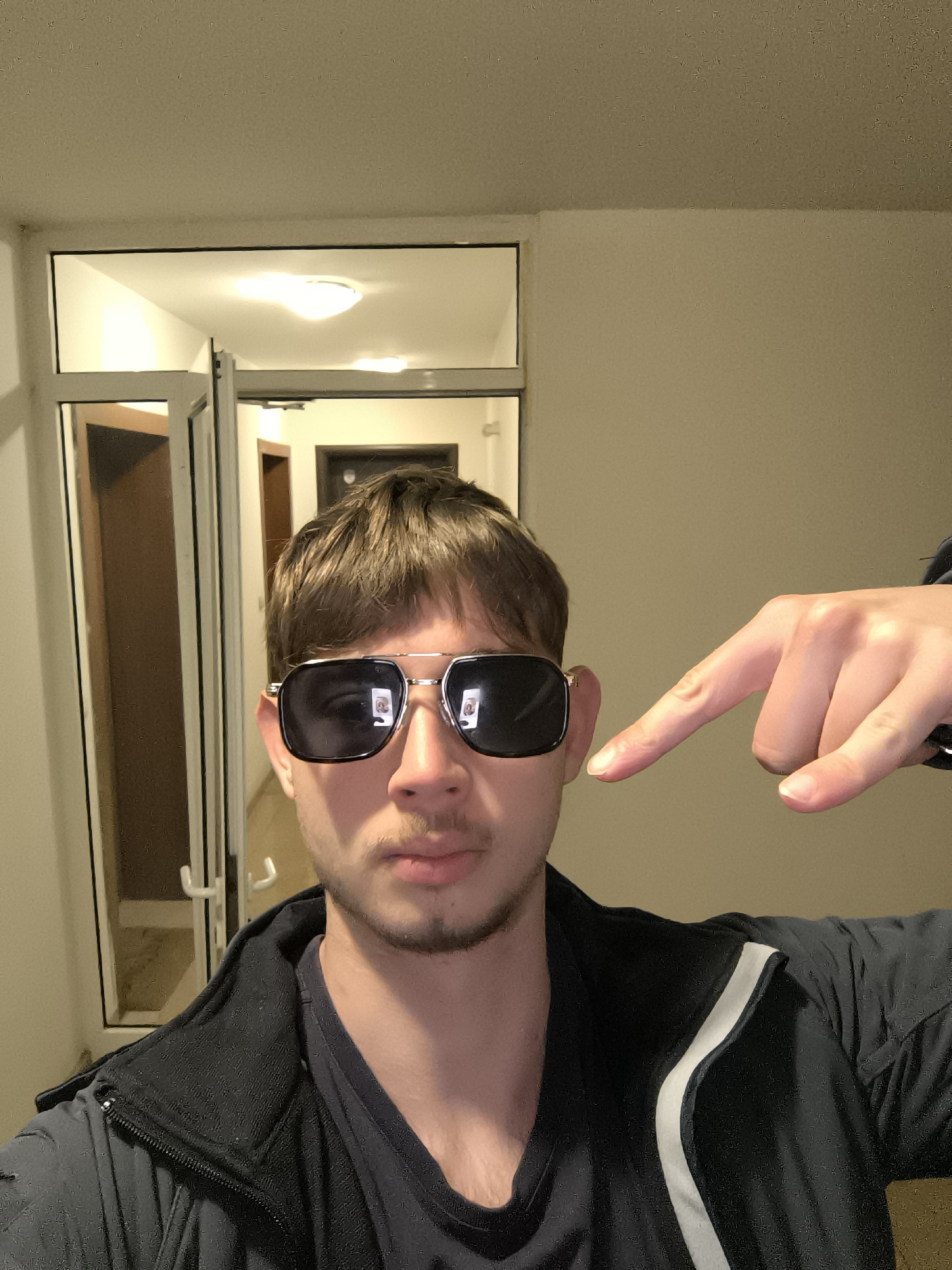 A person wearing sunglasses and a black jacket with white stripes is pointing at their ear. They appear to be standing indoors, possibly in a hallway with a doorway visible in the background. The location is indeterminable, as the provided image doesn't include any features which would allow pinpointing its location.