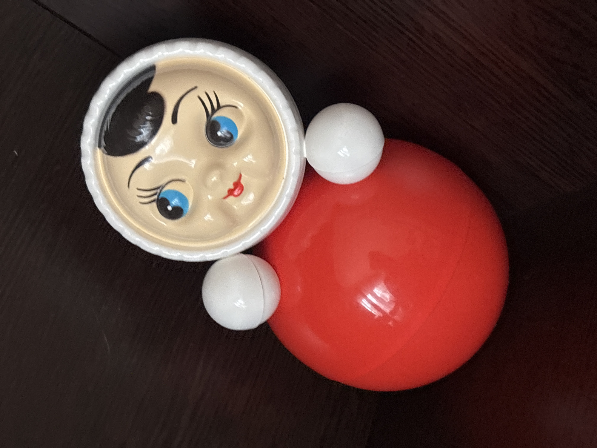A photograph shows a children's roly-poly toy on a dark, wood-grain surface. The toy's head is round and white with a beige face, cartoonish blue eyes, black eyebrows, a black hair curl on its forehead, and a red painted smile. Two small, white spheres protrude from either side of the head, acting as decorative features that resemble ears. The body is a large, red sphere. The object has a glossy texture, reflecting light and suggesting it is made of plastic. The object is a roly-poly toy. The geographic location is unknown.