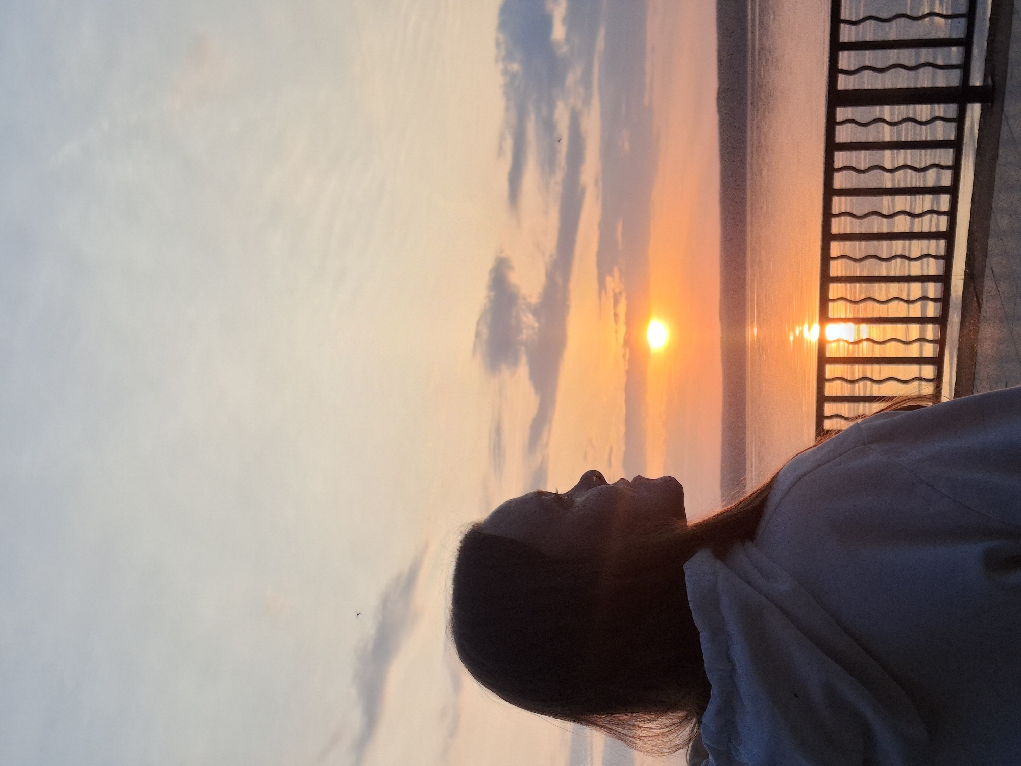 A woman is looking upwards in the evening at the sun reflecting on the water, likely at sunset. The location may be Chaikovsky, Russia, due to the presence of water, possibly a river or lake, and an observation platform or railing. The woman's face is partially silhouetted, and she is wearing a light-colored garment. The sky is a gradient of light blue, yellow, and orange due to the sunset. There are also some clouds. A dark metal railing is on the right side of the image.
