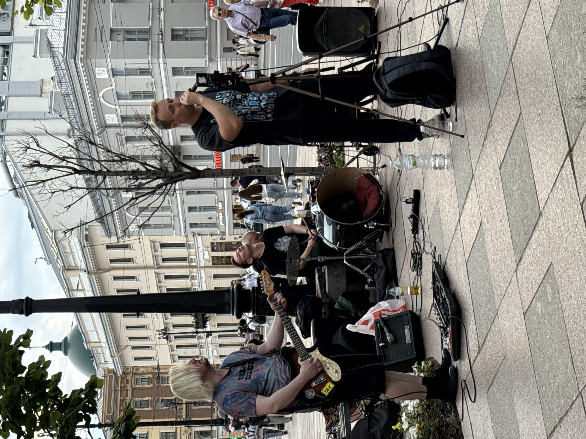 The image depicts an outdoor street performance in Saint Petersburg, Russia. In the foreground, a blonde-haired man is playing an electric guitar. To his right, a man with dark hair is seated behind a drum set. A third man, with blonde hair, stands at a microphone on a tripod, singing. Behind the performers, several buildings with classical architecture are visible, characteristic of Saint Petersburg's cityscape. The ground is paved with large, square stone tiles. Other objects in the scene include musical equipment like amplifiers and a mixing board, as well as a backpack and bottles of water. Pedestrians are present in the background, suggesting a public urban setting.