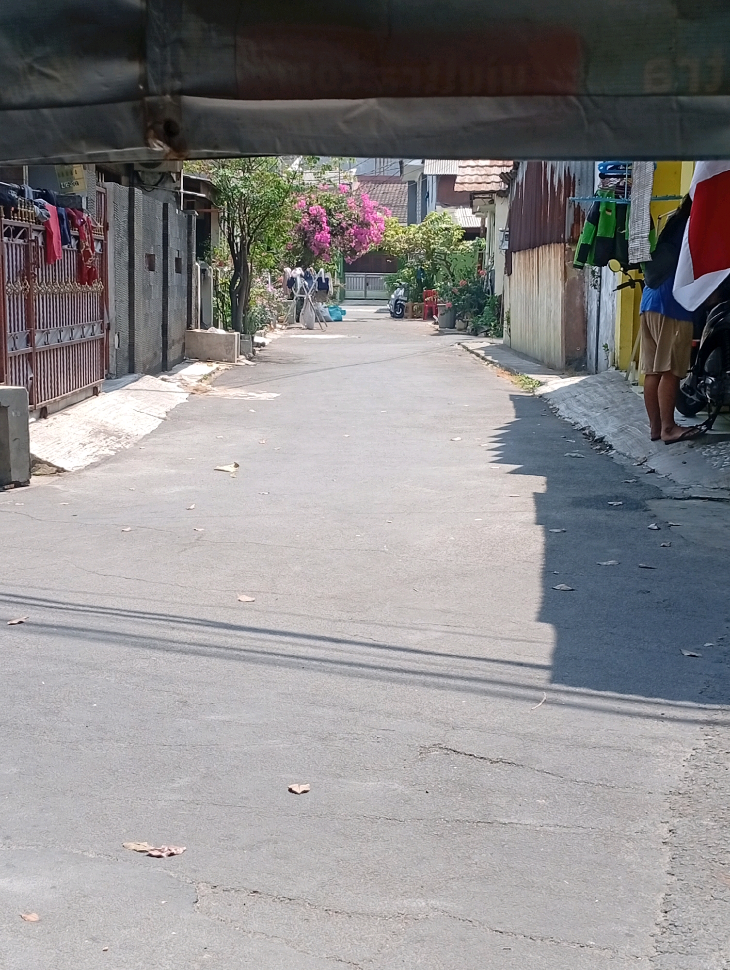 The image shows a narrow, paved street in a residential area of Bekasi, Indonesia. The street is lined with buildings on both sides, some of which appear to be homes with corrugated metal roofs and brick walls. There are several motorcycles parked along the sides of the street, and clothes are hung out to dry on a clothesline in front of one of the buildings. A person is partially visible on the right side of the image, wearing shorts and standing next to a motorcycle. The street itself has some cracks and is littered with a few dry leaves. The scene is illuminated by bright sunlight, casting shadows on the pavement. In the distance, more buildings and vegetation can be seen.