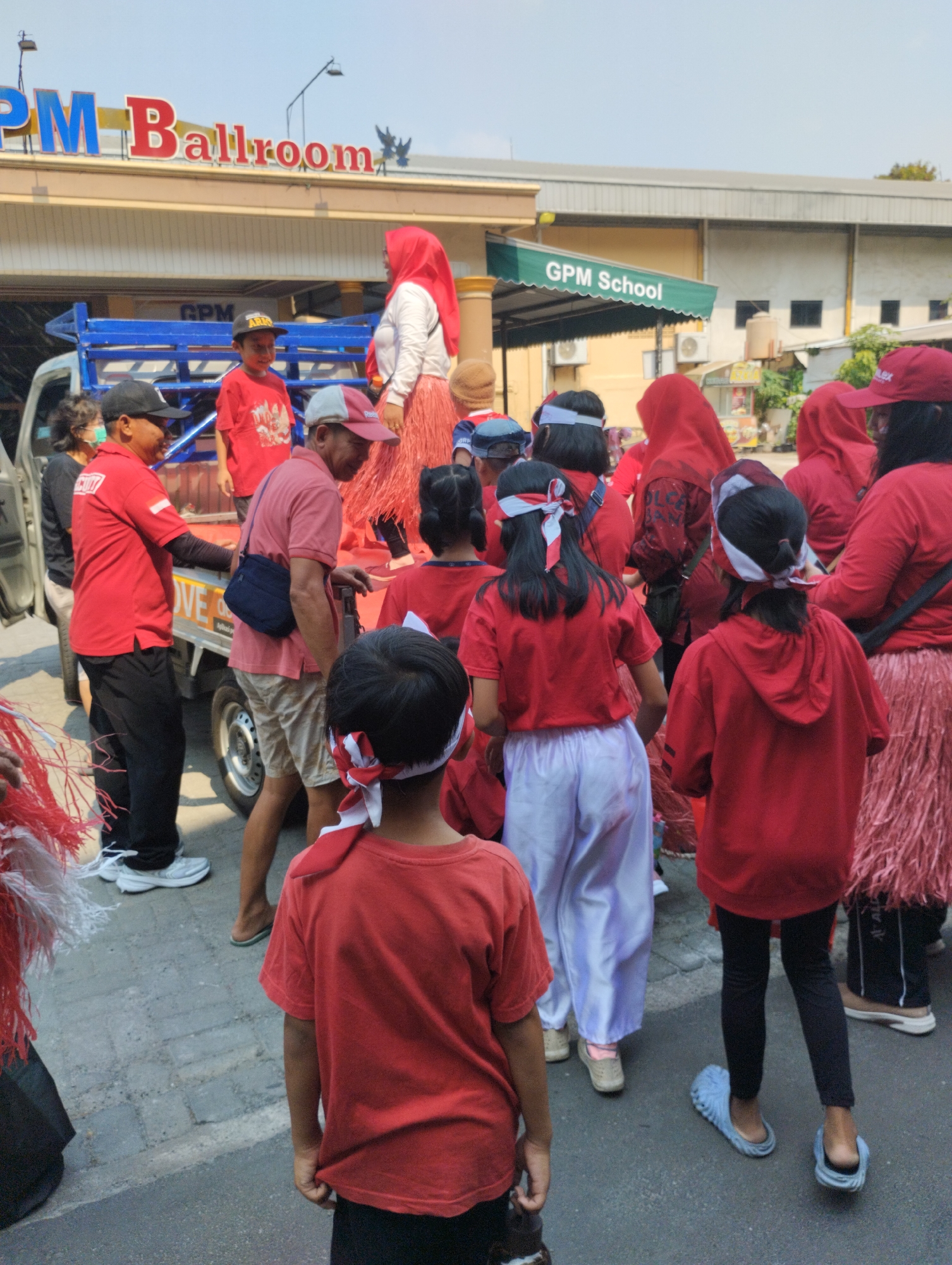 A group of people, many wearing red and white, are gathered outdoors in front of buildings identified as "GPM Ballroom" and "GPM School" in Bekasi, Indonesia. The scene appears to be a street or parking lot with a truck in the foreground. Several children and adults are present, with some of the children wearing headbands with red and white stripes. The people are dressed in a mix of casual attire and what might be ceremonial outfits. One person in the foreground is wearing a red t-shirt, and others are in red shirts, red headscarves, or red skirts. The ground is paved with cobblestones. The buildings in the background suggest an urban or commercial setting. The lighting indicates it is daytime.