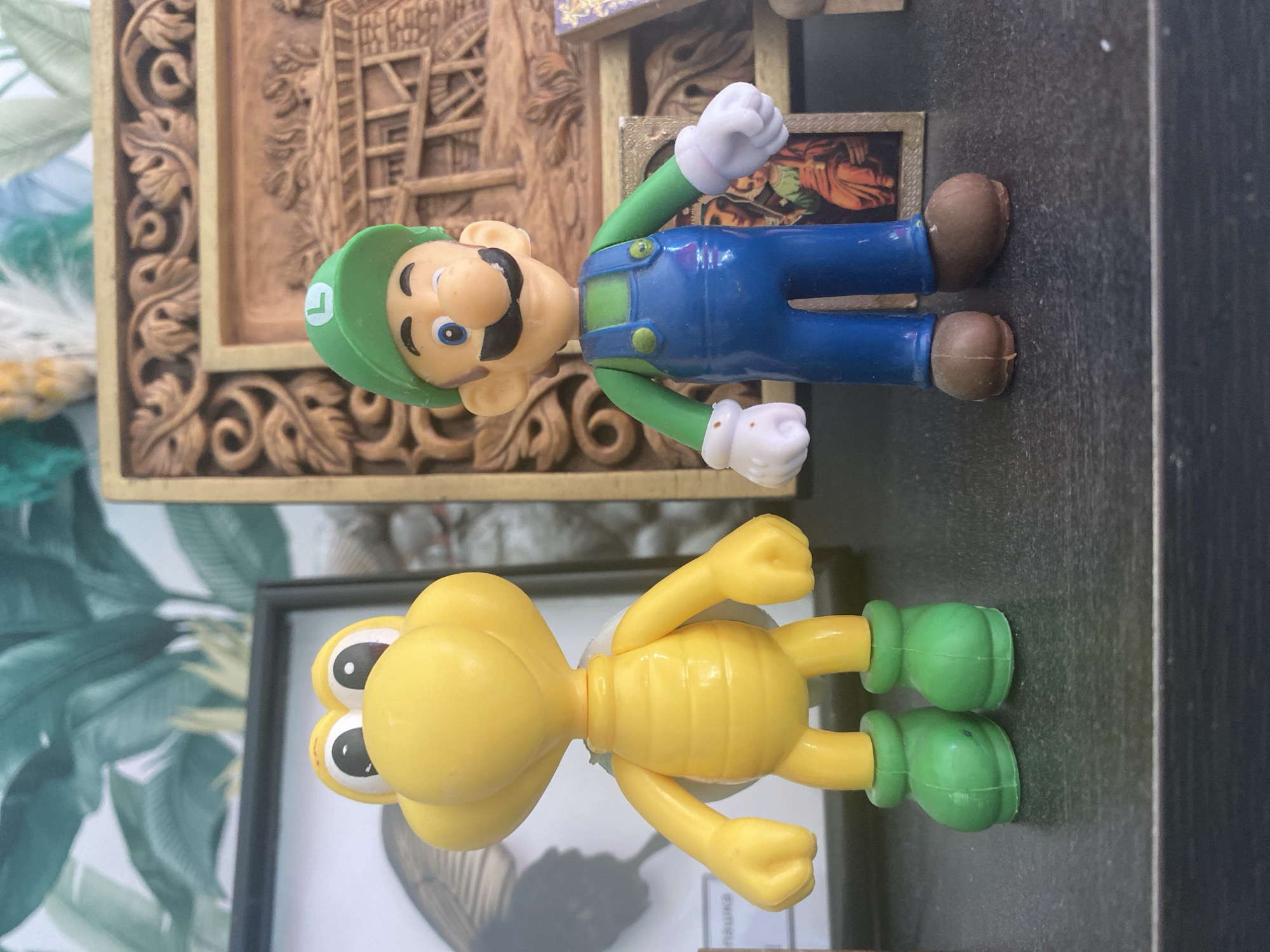 The image shows two toy figurines on a dark, textured surface, likely a shelf. On the left is a yellow Koopa Troopa figure with large eyes, a shell on its back, and green boots. To its right is a Luigi figurine from the Super Mario series, wearing his signature green hat, blue overalls, and red shirt, with white gloves and brown shoes. In the background, a framed piece of art with intricate carvings is visible, along with a tropical-themed wallpaper. There is no indication of the city of Blagoevgrad, Bulgaria, in the image.