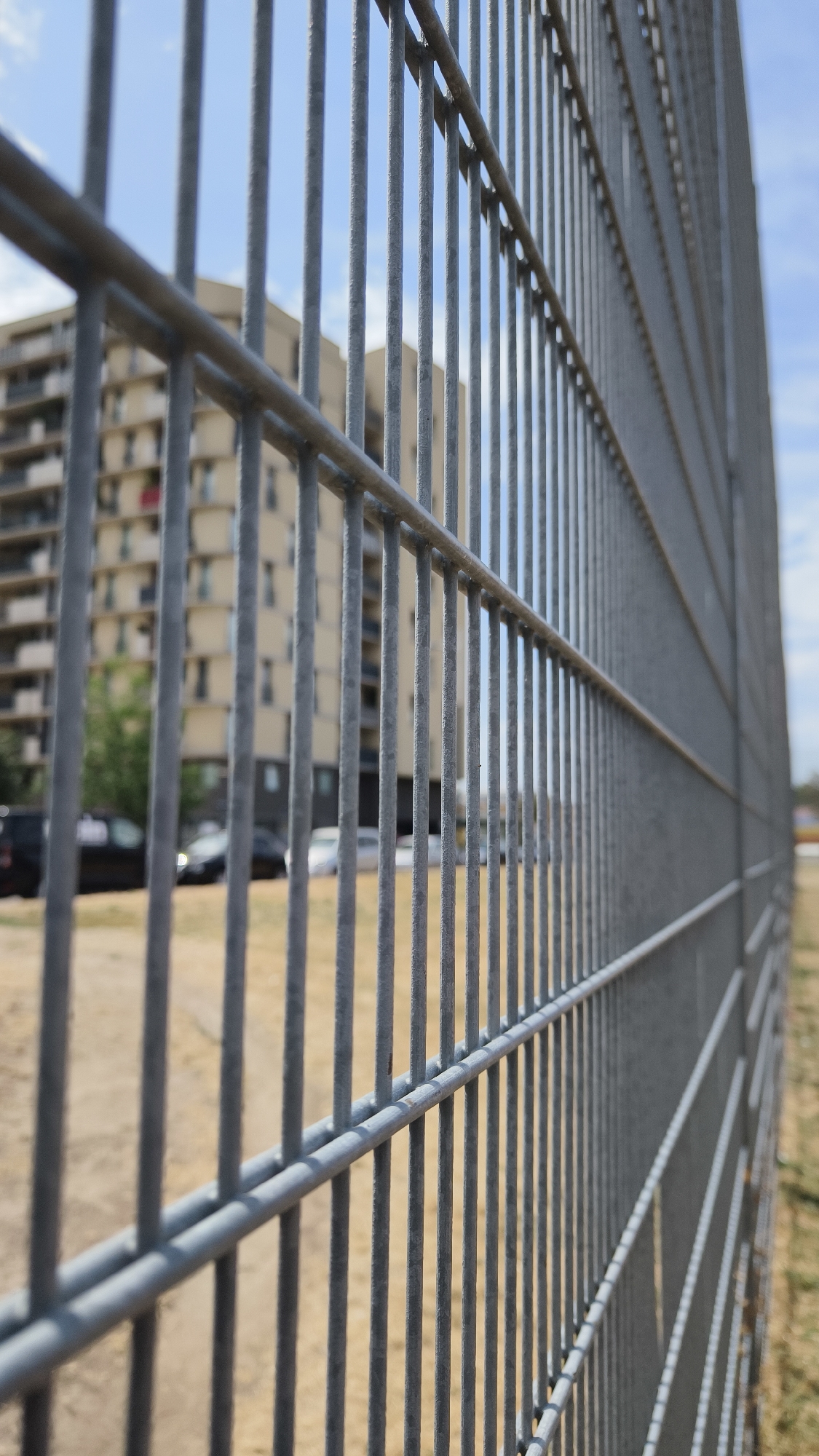 The image is a close-up, eye-level, outdoor shot of a grey metal mesh fence in the foreground, running diagonally from the lower left to the upper right corner of the frame. The fence is composed of vertical bars and horizontal support rails. Beyond the fence, the background is blurred due to a shallow depth of field. Visible in the background is a multi-story, light-colored building with a repeating pattern of windows and balconies. To the left of the building, there are several parked cars and some sparse green foliage. The ground in the background appears to be a dirt or gravel surface. The sky above is partly cloudy with a light blue hue. The scene is static and appears to be a section of a city environment, possibly a park, residential area, or an industrial zone, and is not specifically identifiable as being in Vienna, Austria without further information.