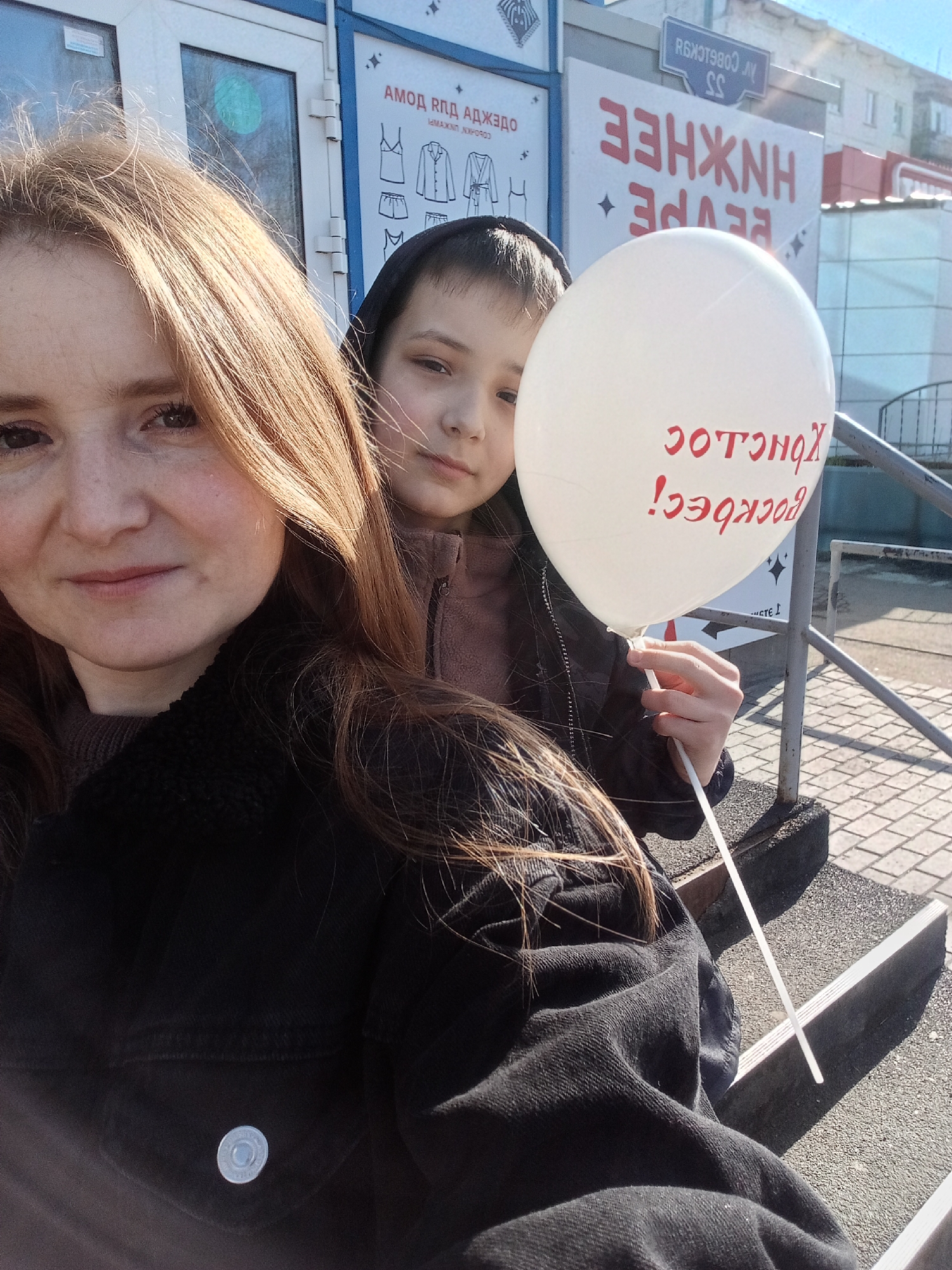 Here is a concise, forensic description of the image:
Two people, likely a mother and child, are posing in front of a building on Sovetskoy Street, in Unknown, Unknown. The woman is in the foreground, her head and shoulders filling the left side of the frame. She has fair skin and reddish-blonde hair. She is wearing a dark jacket. Behind her and to the right is a younger person, possibly a child, with fair skin and dark hair under a hood. They are partially obscured by a white balloon with red reversed (rotated 180 degrees) Cyrillic text which includes "Воскреси" and "Хонсдос." The balloon is held by the child.
The backdrop includes a building with a sign that includes "АМОД ВПД АДЖЭдо" and illustrations of clothing. Above that is a street sign "СОВЕТСКОЙ 55." To the right are some building features, including what appears to be a metal handrail and paved steps. The overall impression is of a candid outdoor shot.