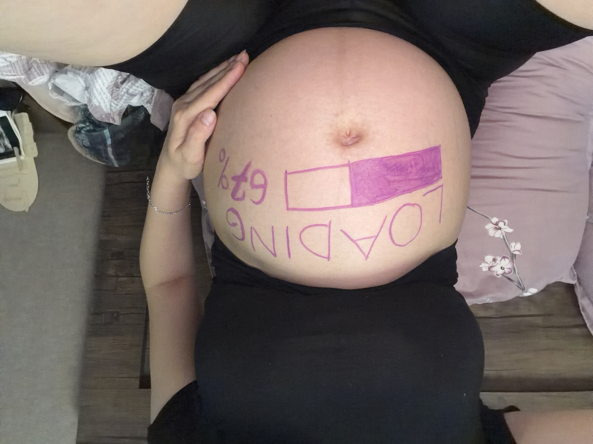 The image shows a close-up of a pregnant woman's belly, which has "LOADING 67%" written on it in pink marker, along with a loading bar graphic. The woman is wearing a black t-shirt and black shorts. She is lying on a bed with pillows, and a blanket is visible in the foreground. The background shows a wooden headboard and a wall. The scene appears to be indoors, in a domestic setting, and there is no information to suggest it is in Tbilisi, Georgia.