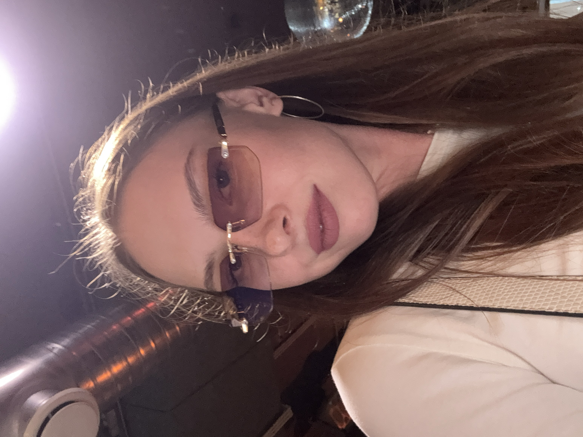 A medium shot shows a young woman with long, brown hair and light skin. She is wearing large, square, rimless sunglasses with a pinkish tint and large silver hoop earrings. Her lips are painted with a matte pink lipstick. She is wearing a light-colored top with some texture visible on the strap of what appears to be a purse. The background is dark, with a metal pipe and a light fixture visible on the upper left, and a blurry, reflective object that may be a disco ball on the right. The scene appears to be indoors, possibly in a bar or club. There is no information to suggest this location is in Perm, Russia.