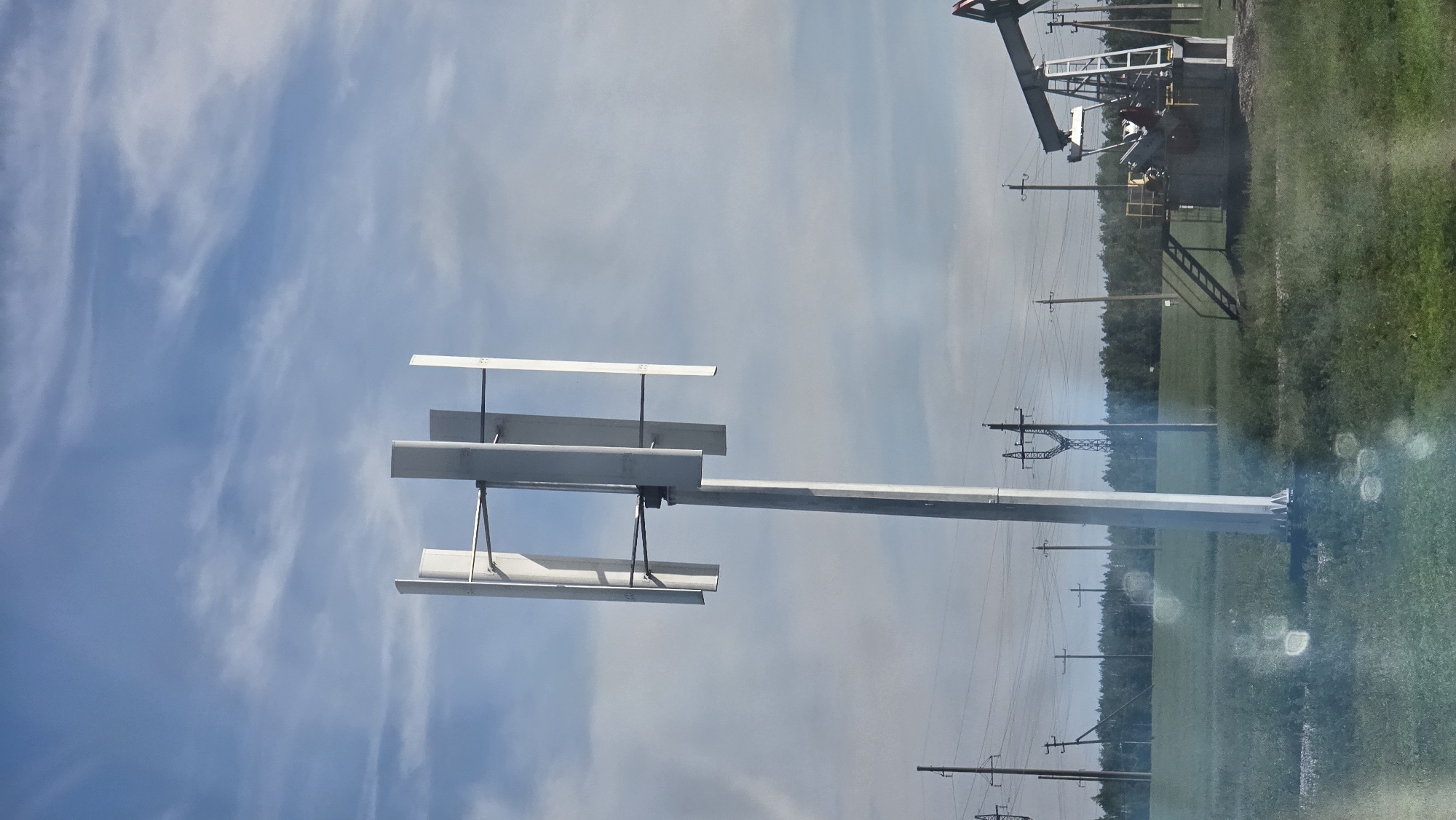 In an open field under a cloudy blue sky, a vertical axis wind turbine stands tall. The turbine features three rectangular blades arranged vertically on a central pole, with supporting horizontal rods. To the right, a pumpjack, characteristic of an oil extraction site, is visible. The landscape is dotted with power lines and poles, indicating a rural or industrial setting. A dense line of trees forms the horizon in the background. The foreground is a green field with some areas of dry grass and a few circular light flares.