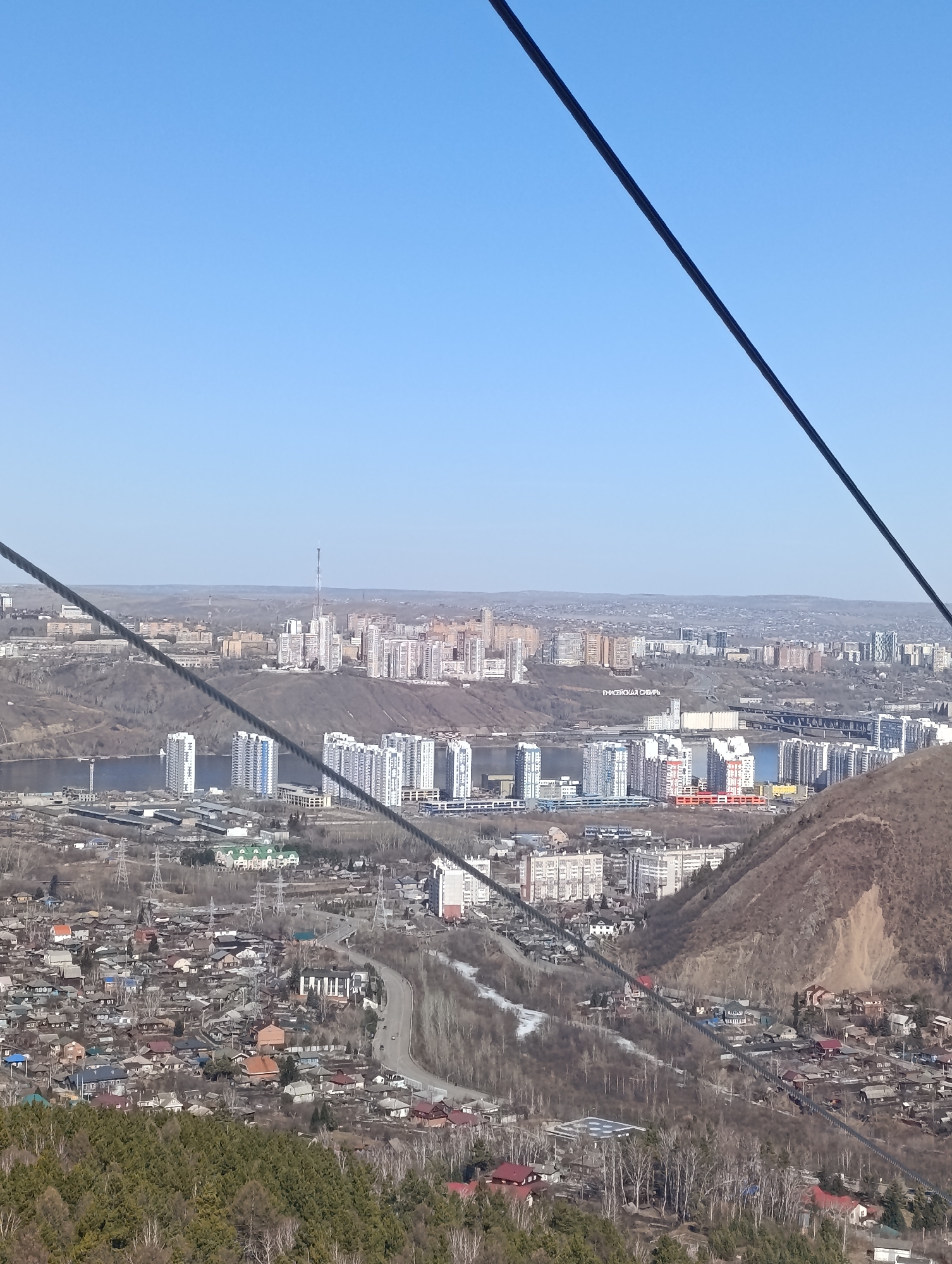 The image shows a view of Krasnoyarsk, Russia from a high vantage point, possibly a cable car. In the foreground are dense clusters of residential homes and buildings, some with distinct red roofs, interspersed with bare trees. A winding road cuts through the built-up area. In the middle ground, a wide river separates the foreground settlement from a city skyline dominated by high-rise apartment buildings. A large white inscription reading "Енисейская Сибирь" (Yenisey Siberia) is visible on a hillside. Further back, the urban sprawl continues to the horizon beneath a clear blue sky. A few cable lines stretch across the top portion of the image, suggesting the shot was taken from a cable car.