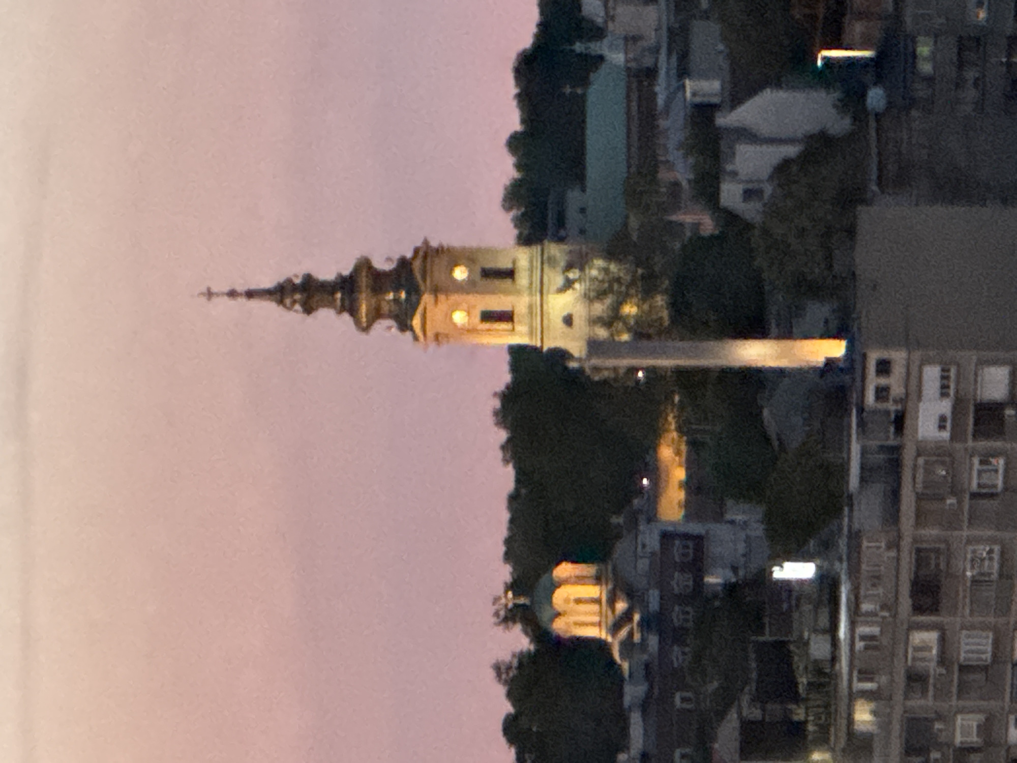 The image shows an evening cityscape featuring the tower of the Cathedral Church of St. Michael the Archangel in Belgrade, Serbia. The tower is illuminated, with its ornate spire pointing upward. Nearby, a building with a green dome is also visible, lit up against the dusk sky. Modern apartment blocks stand in the background, alongside other buildings and trees. The sky is a soft pink hue, suggesting either sunset or twilight. There are no people visible in the image.