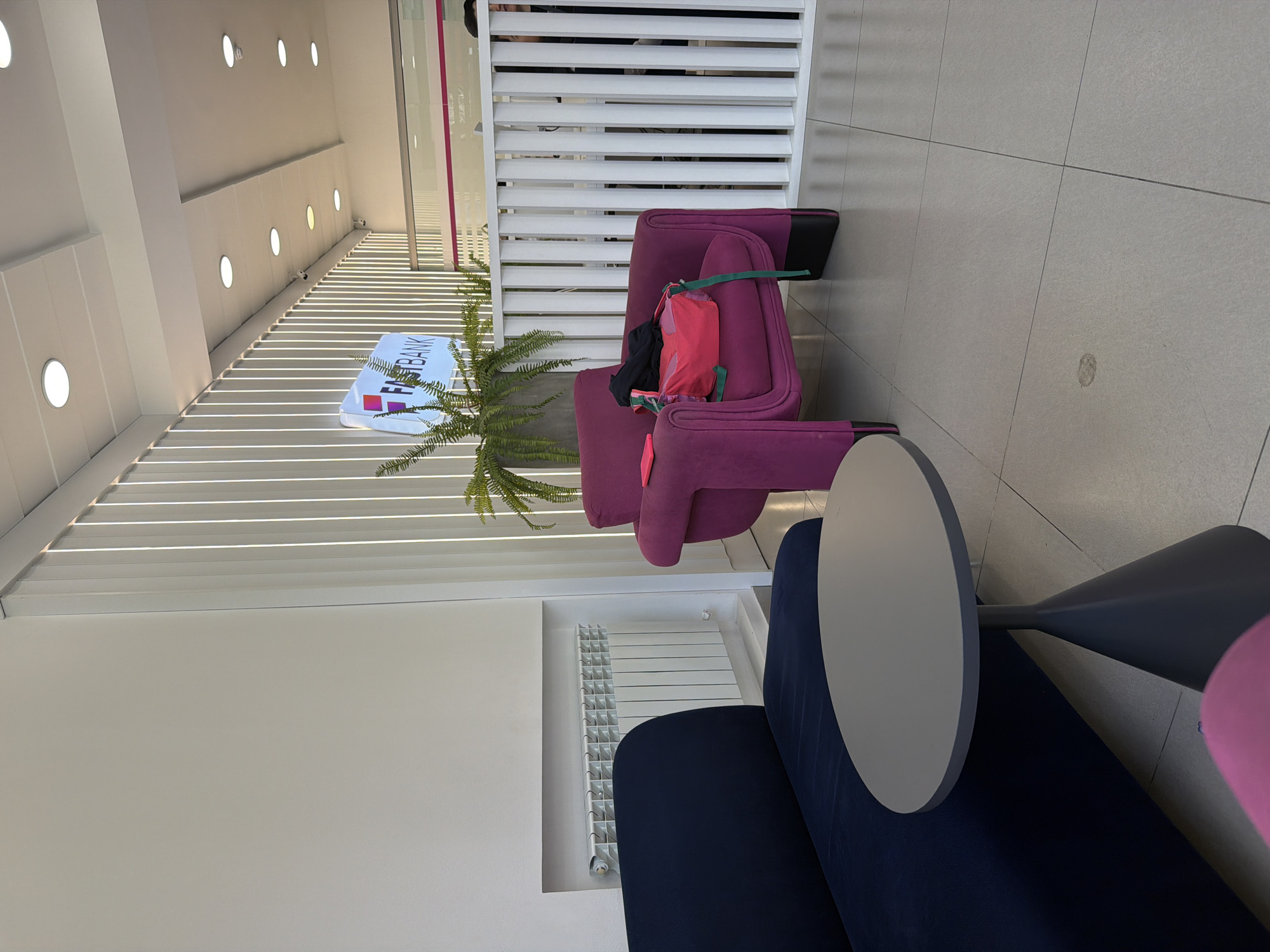 The image captures an interior setting in Yerevan, Armenia, featuring a modern waiting area within a facility, possibly a bank. The area is arranged with purple upholstered chairs and a circular table with a grey tabletop and a dark grey base, placed next to a navy-blue upholstered sofa. A leafy green plant sits between the chairs and a white slatted partition wall. The setting also includes a white radiator installed in the wall behind the seating area. The presence of the "FAST BANK" logo suggests the location is within a branch of the bank. The interior is characterized by a clean, modern aesthetic with an emphasis on comfort for waiting patrons.