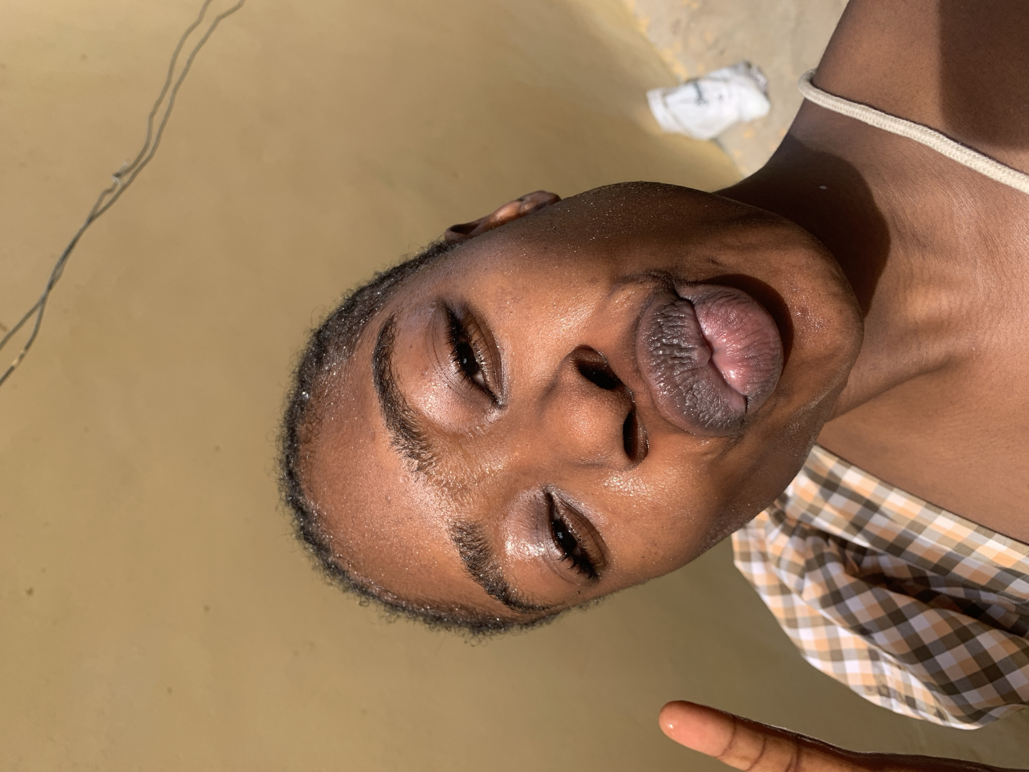A close-up, selfie-style portrait shows a young Black woman with wet skin, likely from sweat or water. She is looking at the camera with her lips pursed in a kissing gesture. Her right eye is partially closed, and she is raising her left index finger towards the camera in a peace sign. The background is a textured, yellow wall. The woman is wearing a checkered top with orange, brown, and white squares. A white strap from her clothing is visible on her right shoulder. In the upper right background, a white object, possibly a piece of clothing or paper, lies on the ground, which appears to be a dirt or concrete surface. There are some wires running across the top of the background. The lighting suggests it is daytime, and the scene is outdoors, likely in Rafin Par, Nigeria.