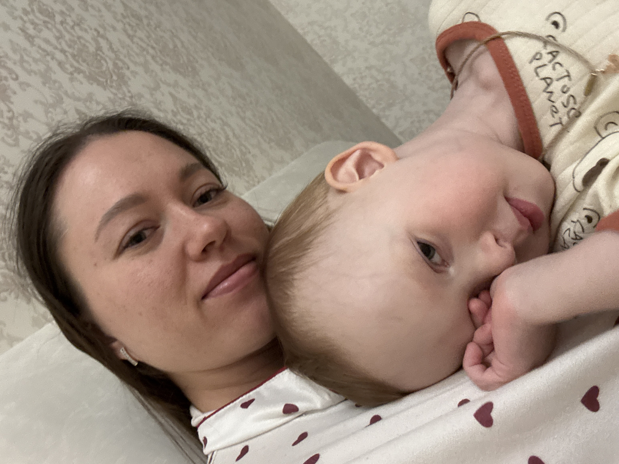 A medium-shot captures a woman and child lying in what appears to be a domestic setting.
The woman, on the left, has dark hair and fair skin, wearing what appears to be patterned pajamas. She is angled towards the camera, engaging the viewer with a soft smile.
A fair-skinned baby lies beside her, possibly resting on the woman's chest. The baby is dressed in pajamas, with the wording "CACTUS PLANET" visible on the fabric. The baby's face is turned towards the camera, with one hand near their mouth.
The setting seems to be indoors, likely a bedroom. A patterned wall is visible in the background, and the bed is covered in a patterned sheet.
The location is presumed to be in the city of Ферма, Russia.