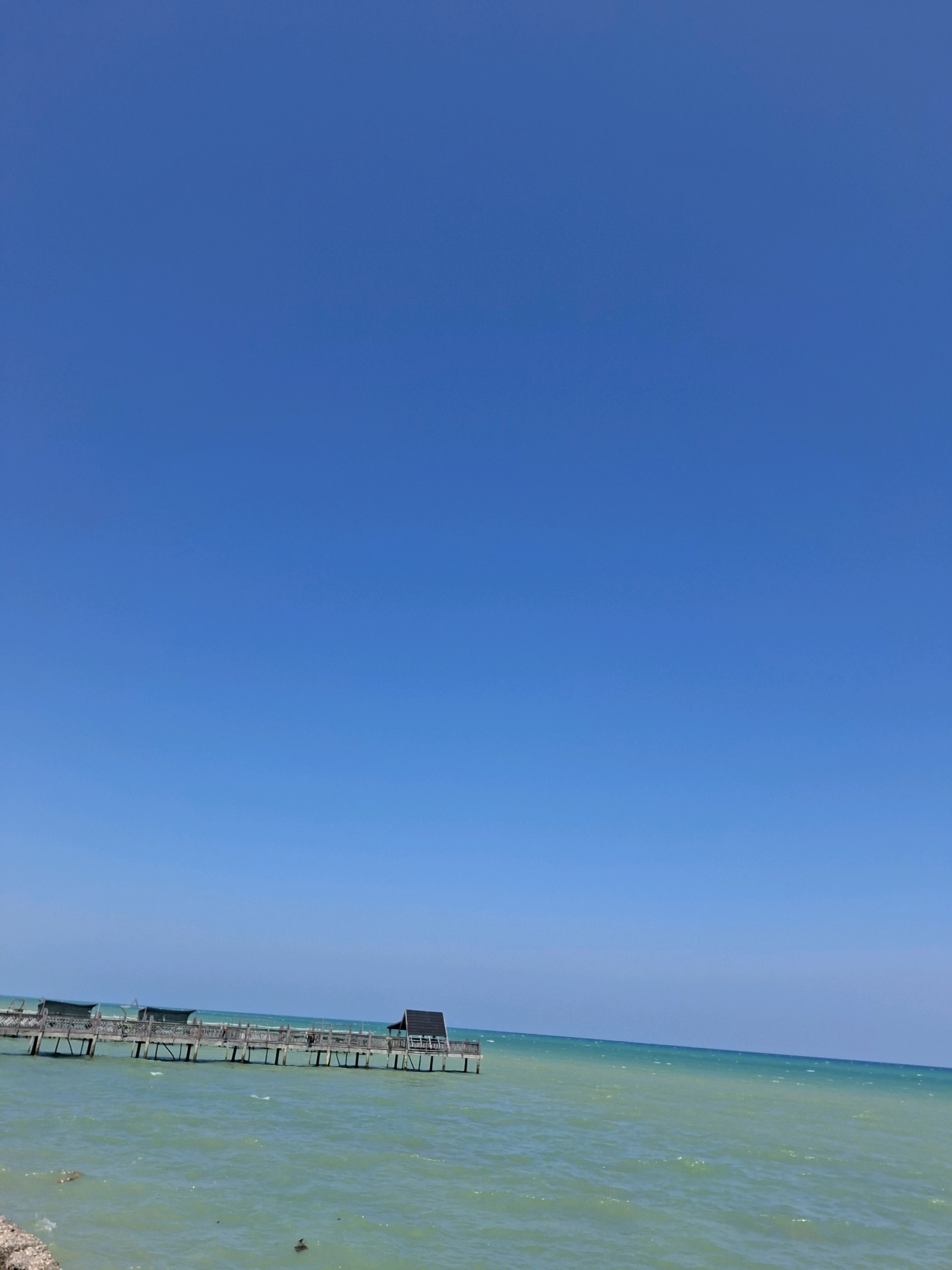 A wooden pier extends into a turquoise body of water under a clear blue sky. The pier, constructed with weathered planks and supported by pilings, features several covered sections and a small, dark-roofed structure at its end. The water is slightly choppy with visible ripples. The horizon is visible in the distance where the water meets the sky. The scene appears to be a coastal area in Sejati, Indonesia.