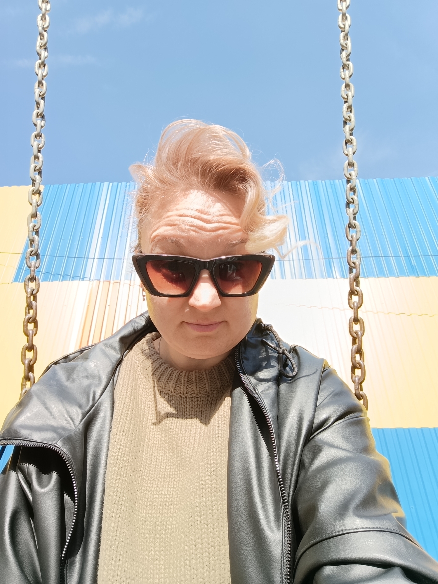 A low-angle portrait depicts an adult female with light blonde hair and dark, cat-eye sunglasses. She wears a dark jacket over an olive-toned ribbed knit sweater. Two heavy metal chains, consistent with a swing, flank the subject on either side, extending from the top of the frame. The background consists of a clear blue sky overlying a structure composed of vertical corrugated metal panels painted in alternating blue and yellow stripes. The scene is located in Chaikovsky, Russia.
