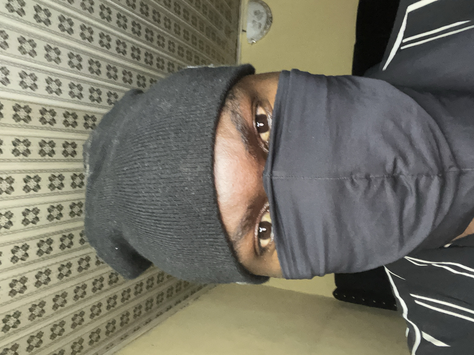A close-up portrait captures a person from the shoulders up in an indoor setting. The individual has dark skin and prominent dark brown eyes, which are the main visible facial features. The lower part of their face, including the nose and mouth, is covered by a dark, possibly black, fabric, resembling a balaclava or a pulled-up gaiter. They are also wearing a dark grey knitted beanie on their head, fully covering their hair and ears.
On the right side of the image, a portion of a dark garment, likely a jacket or shirt, is visible, featuring distinct white horizontal stripes. The background consists of a wall with a repeating pattern of light grey horizontal stripes, each adorned with rows of dark floral or geometric motifs. Above the person's head, affixed to the wall, is a small, light-colored, semicircular object, possibly a light fixture or decorative element. The lower portion of the image shows a plain, light-colored surface, likely a floor.
The person is looking directly into the camera, with the eyes appearing focused and neutral. Catchlights are visible in their pupils, suggesting a light source in front. There is no specific activity depicted other than posing for the photograph. The overall mood conveyed by the concealed face and direct gaze is somewhat enigmatic. No text is visible in the image. The location context provided for this image is Gura Top, Nigeria.