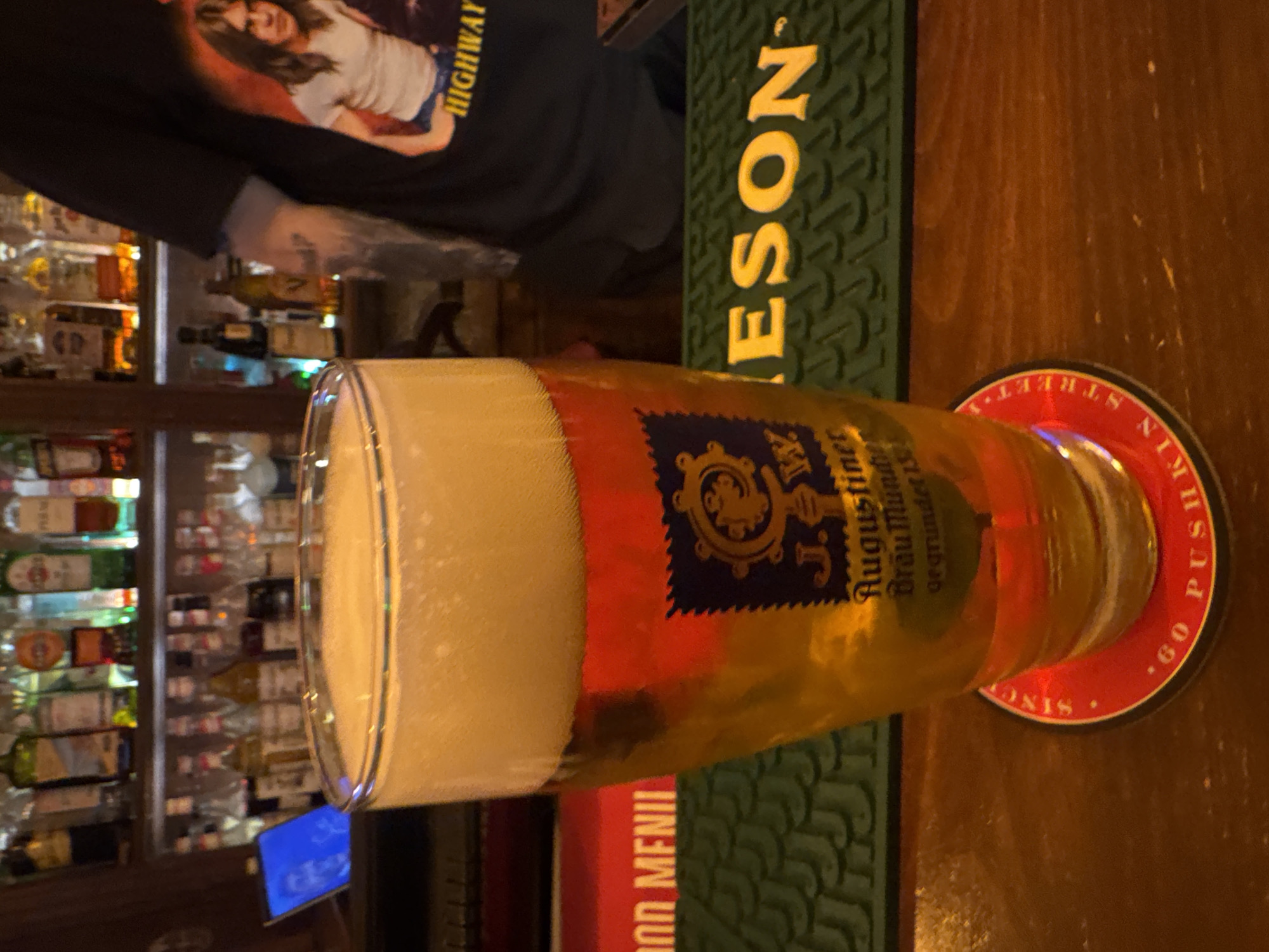 The image shows a glass of Augustiner beer sitting on a red coaster with white lettering, resting on a wooden table in a bar. The glass is tall and filled with beer, featuring a large head of foam. The label on the glass reads "J. Augustiner Brau Munchen." A green Jameson bar mat is also visible next to the beer glass. A person wearing a black t-shirt with a graphic of a woman on it is standing nearby. Behind the beer, the background includes a well-stocked bar with numerous liquor bottles displayed on shelves. A blue-lit menu screen is visible as well. The location appears to be a pub or bar, but there is no indication of it being in Yerevan, Armenia.