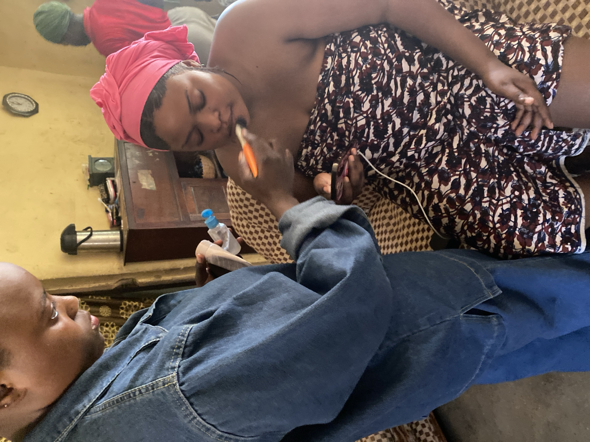 In Panyam, Nigeria, an individual wearing a denim jacket is applying makeup to another person who is wrapped in a patterned cloth and wearing a pink headscarf. The makeup application appears to be focused on the lips. The environment seems to be an indoor setting, possibly a home, with furniture and a clock visible in the background. Another person in a red shirt and green headscarf is partially visible in the background.