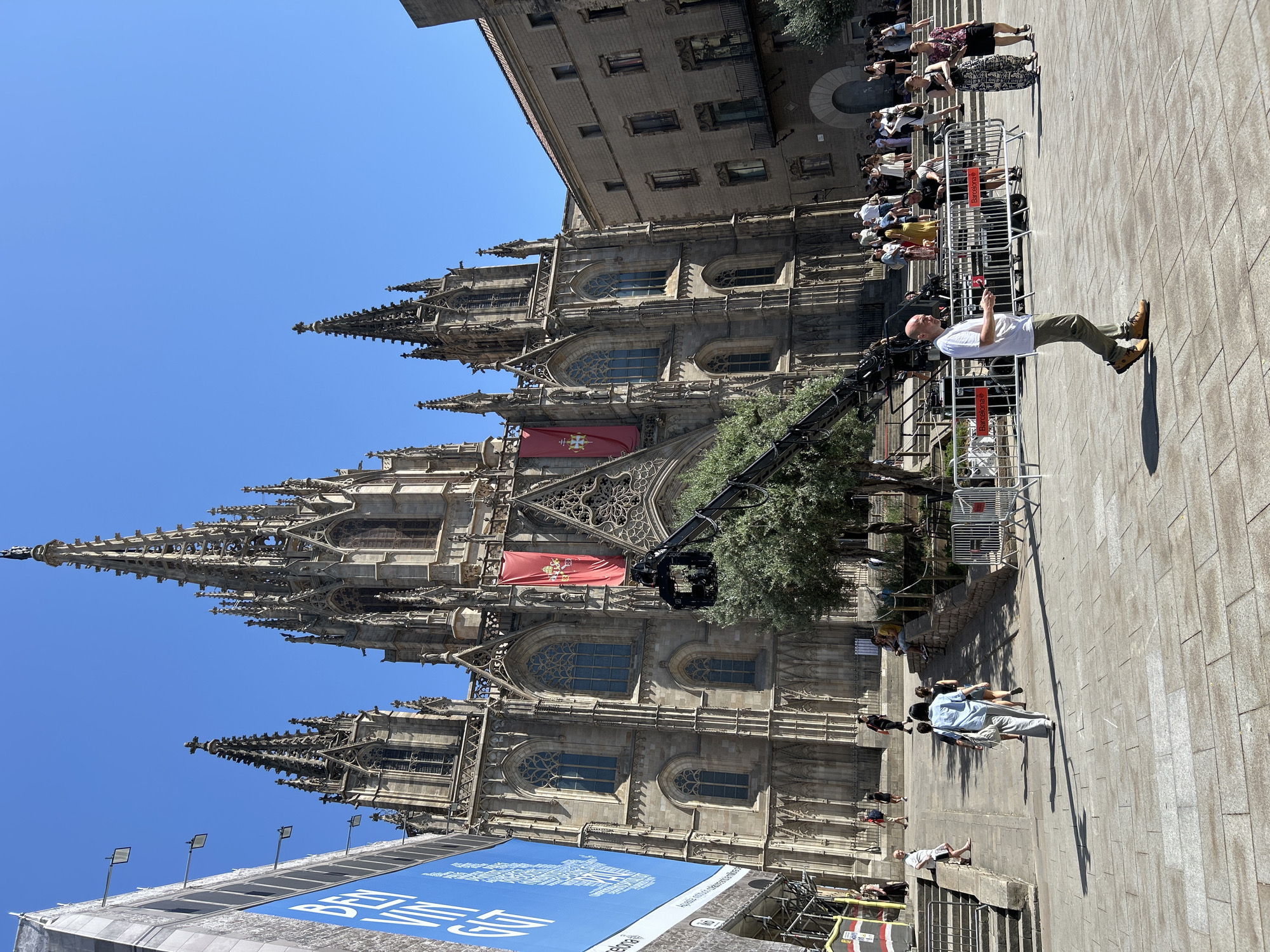 This is a medium-shot, eye-level, outdoor photograph taken on a sunny day in Barcelona, Spain. The main subject is the exterior of a large, ornate Gothic cathedral, identified as the Barcelona Cathedral, with its characteristic spires and intricate stone carvings. Two large red banners with a coat of arms are hanging from the facade. In front of the cathedral, a film crane is set up with a camera mounted on it, suggesting a film or television production is underway. Several people are present on the plaza in front of the cathedral, some walking, others standing in small groups. A man in a white t-shirt and green pants is walking towards the camera in the foreground. The plaza is paved with large stone tiles, and there are metal barricades set up in some areas. To the left of the cathedral, a large blue banner with white text is visible, likely an advertisement or informational display. The sky is clear and blue.