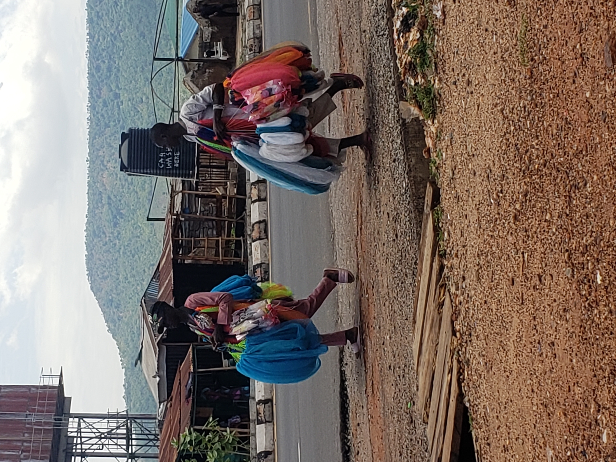 This outdoor image, set in Adankolo, Nigeria, depicts two individuals, likely street vendors, walking along a roadside path during daylight hours under a partly cloudy sky. The scene shows a paved road running parallel to a dirt and gravel path.
Both individuals are carrying large, voluminous bundles of colorful merchandise, possibly textiles or lightweight clothing, on their heads or shoulders. The bundles feature a mix of colors including reds, oranges, pinks, blues, whites, and greens. The first person, closer to the viewer, wears a light-colored long-sleeved shirt and dark lower garments, with sandals. The second person, slightly further away, is dressed in a reddish-brown top and dark trousers, also wearing sandals and carrying a similar load.
In the background, on the left side of the road, various structures are present, including buildings made with corrugated metal sheets. A prominent black plastic water tank is visible, with partially legible text that reads "CAR WASH HERE". Beyond these structures, a range of large, vegetated hills or mountains stretches across the horizon. The setting suggests a semi-urban or rural environment, characterized by basic infrastructure alongside natural landscapes. Sparse green vegetation can be seen growing along the edges of the dirt path. On the far left, a structure resembling scaffolding or an incomplete building is also visible.