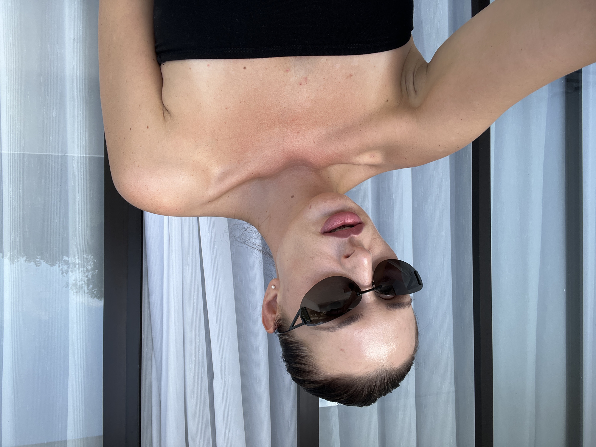 A woman wearing sunglasses and a black top is photographed indoors against a backdrop of white curtains. The photograph has been rotated, presenting an upside-down view. The woman's skin appears slightly sunburned across the shoulders and chest. The location is Kuta Selatan, Indonesia.