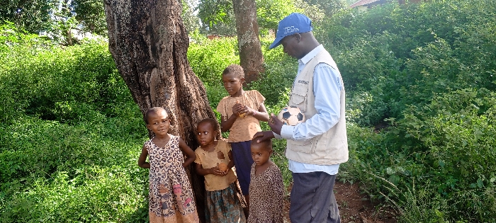 The scene is set outdoors in Ngalula, Democratic Republic of the Congo, against a backdrop of dense green foliage and a large, dark-barked tree trunk on the left. In the distance, a partial reddish-brown roof of a structure is visible above the vegetation. Five individuals are present: one adult male and four children.
The adult male, positioned to the right of the group, wears a light blue long-sleeved shirt, a beige multi-pocket vest, and a blue baseball cap with white lettering. He is standing, facing the children, and holds a small, black and white checkered soccer ball in his hands. His left hand is placed on the head of the smallest child.
Four children are clustered to the left of the adult, near the tree trunk. From left to right:
1. A young girl in a light-colored dress with a dark heart or animal print, looking towards the viewer with a slight smile.
2. A small child in a tattered light brown top and dark bottom, looking towards the adult male.
3. A slightly older child, appearing to be a girl, in a light brown t-shirt and dark trousers, also looking at the adult male and holding a small, brown, spherical object in her hands.
4. The smallest child, in a patterned dress, positioned directly in front of the adult male and looking downwards, with the adult's hand resting on their head.
The interaction involves the adult male presenting the soccer ball to the children, who are gathered around him.