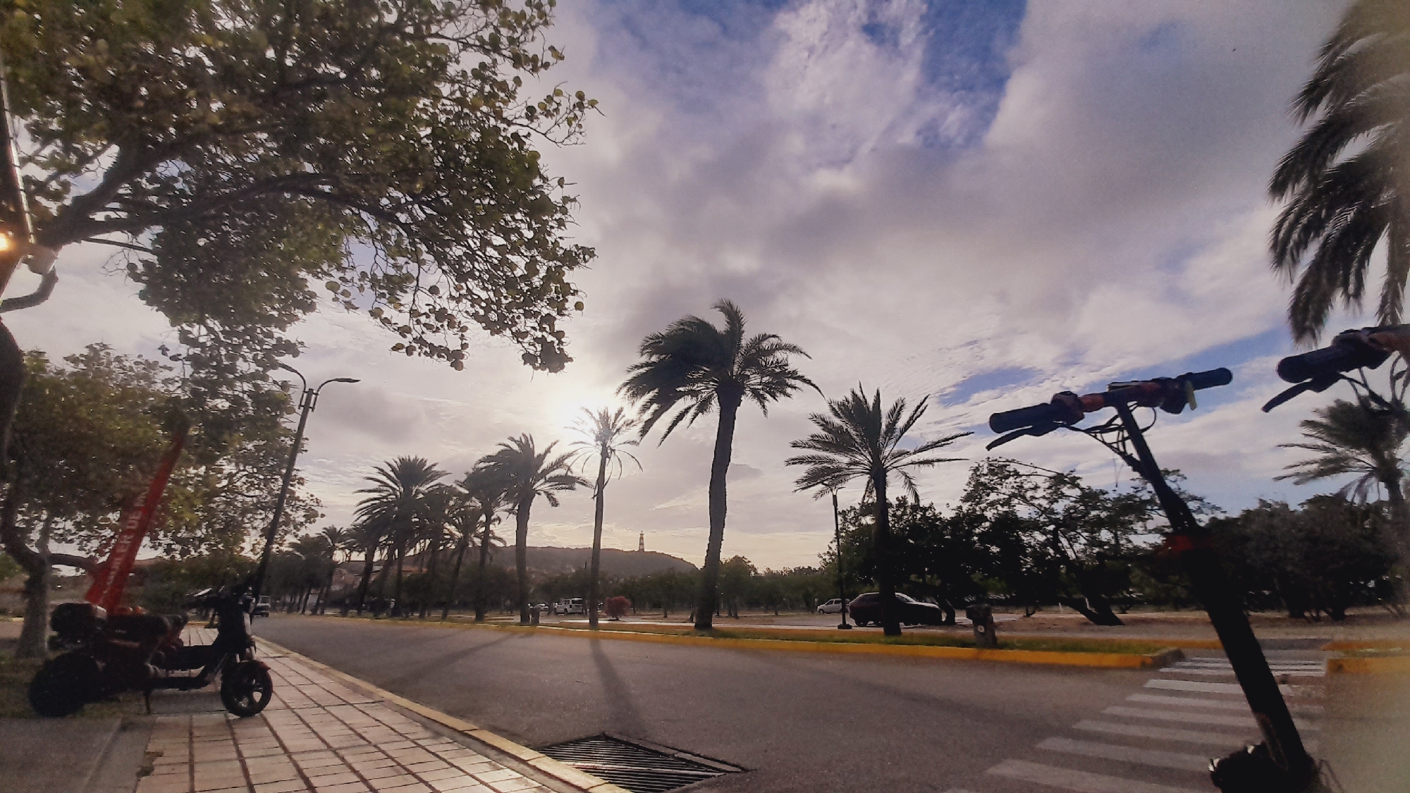 The image depicts a street scene with several palm trees and lush greenery under a cloudy sky with patches of blue. In the foreground, on the right side, two electric scooters are visible, with their handlebars and steering columns dominating the frame. On the left side, another electric scooter is parked on a paved sidewalk alongside a street lamp. The sidewalk is made of rectangular pavers. The street itself is paved and appears to be a two-way road with a yellow line marking the edge. There are some cars parked in the distance and a lighthouse-like structure visible on a hill. The overall impression is a sunny but slightly overcast day in a tropical or Mediterranean-like setting. The city of Pedro González, Venezuela, is known for its beaches and tropical climate, which aligns with the visual elements in the image, suggesting this scene could be from that location.