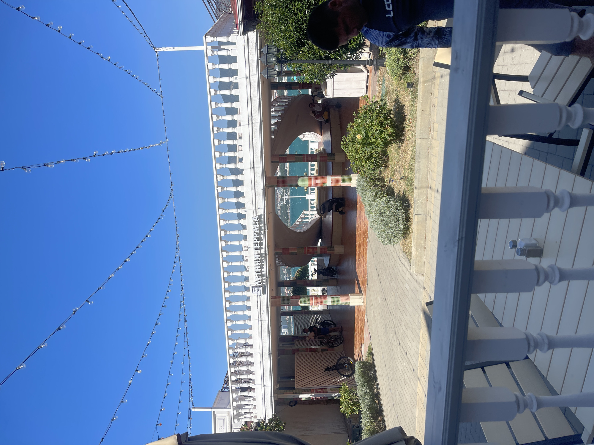 The image shows a bright, sunny day with a clear blue sky over Gelendzhik, Russia. A white, ornate balustrade is visible in the foreground, with a table and salt and pepper shakers on it. Beyond the balustrade, a multi-story structure with a white balcony and balustrade is visible, featuring a spiral staircase leading up to it. The structure appears to be a cafe or restaurant, with the text "ЛЕТНЯЯ ТЕРРАСА" (Summer Terrace) visible on the facade. Several people are present, some sitting on benches or stairs, and others standing. Two bicycles are parked near the entrance to the structure. Lush green bushes and trees are scattered around the area, and a paved pathway is visible. In the background, the sea is visible, with boats on the water, and mountains can be seen in the distance. A man in a dark blue shirt with a graphic design is visible in the lower right corner of the image. The overall scene depicts a pleasant outdoor recreational area in a coastal city.