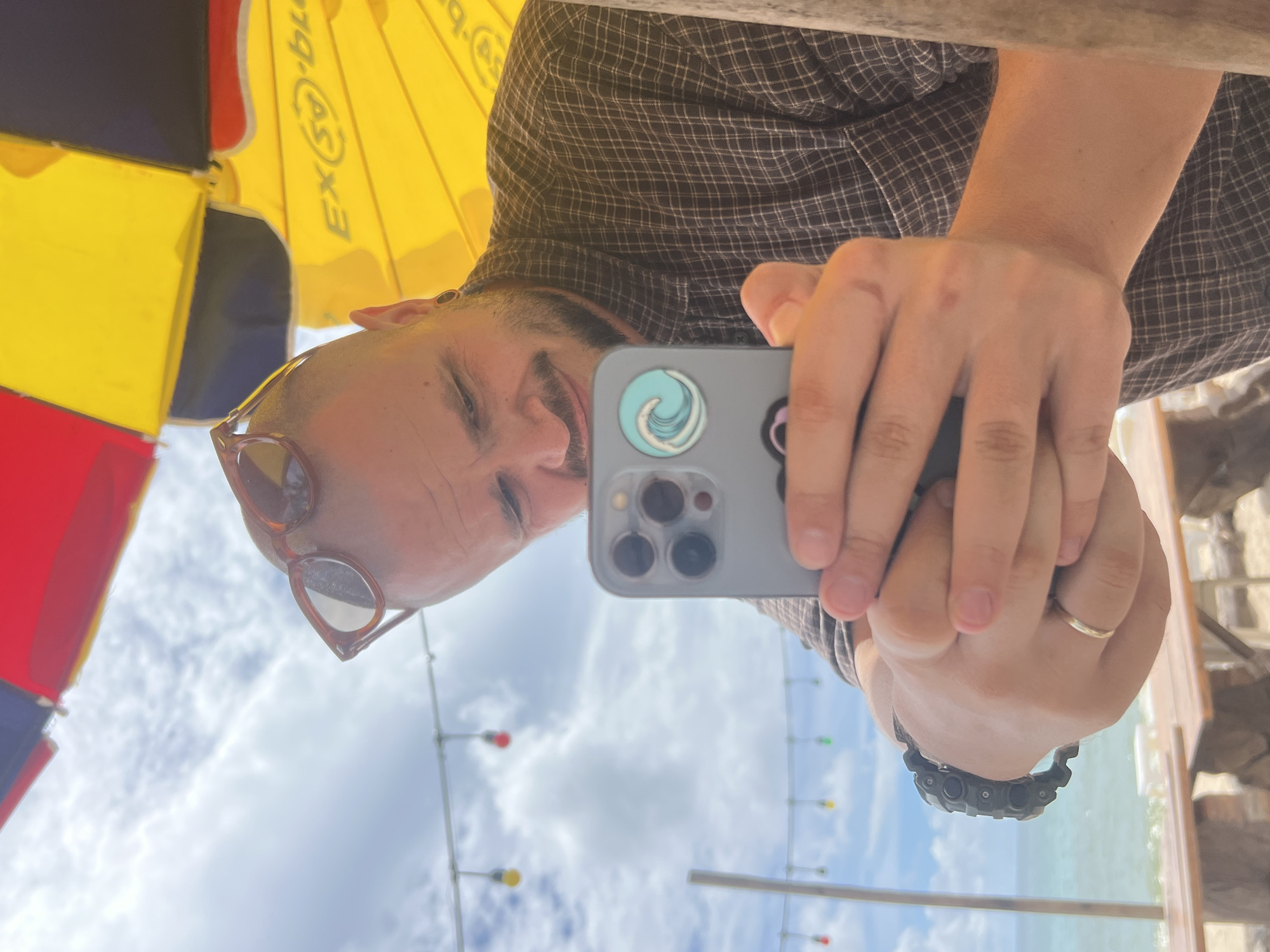 A man, appearing to be in his late 20s or early 30s, with a shaved head, beard, and sunglasses perched on his head, is taking a selfie with his smartphone. He is wearing a dark plaid shirt. He is seated at a wooden table at an outdoor cafe or beachside establishment, with a yellow and red umbrella providing shade above him. The background reveals a cloudy sky and a distant view of the ocean and beach. String lights are visible overhead. The phone case has a circular sticker with a wave design. He is wearing a watch on his left wrist and a ring on his left ring finger. The scene is likely in Choeng Thale, Thailand, given the presence of beachside amenities and tropical scenery.