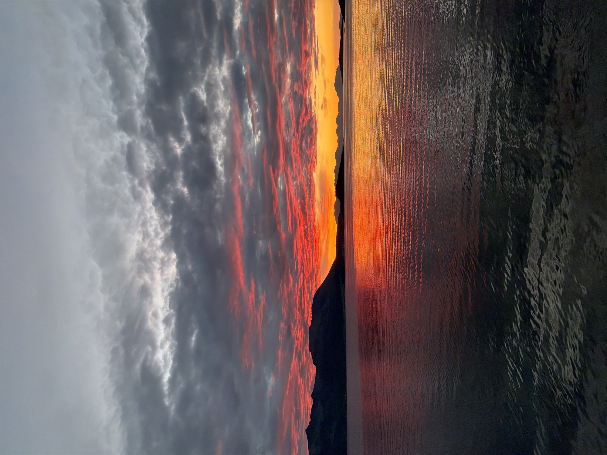 This image captures a dramatic sunset over the waters of Harstad, Norway. The sky is a spectacle of color, with fiery reds and oranges dominating the lower horizon, transitioning to darker grays and muted tones at the zenith. Clouds are densely packed, with the lower layers vividly illuminated by the setting sun, creating a textured, painterly effect.
In the middle ground, a silhouetted mountain range stretches across the scene, its dark peaks sharply defined against the vibrant sky. The water in the foreground reflects the intense colors of the sunset, with the rippling surface creating streaks of light and shadow. The lower portion of the water is darker, with textured patterns of light playing on its surface, suggesting depth and movement.
There are no people or specific man-made structures visible in this panoramic view. The scene is entirely natural, focusing on the interplay of light, water, and landscape. The overall impression is one of natural beauty and the awe-inspiring power of a Nordic sunset. The location is specific to Harstad, Norway, a coastal town known for its stunning natural scenery and dramatic celestial displays.