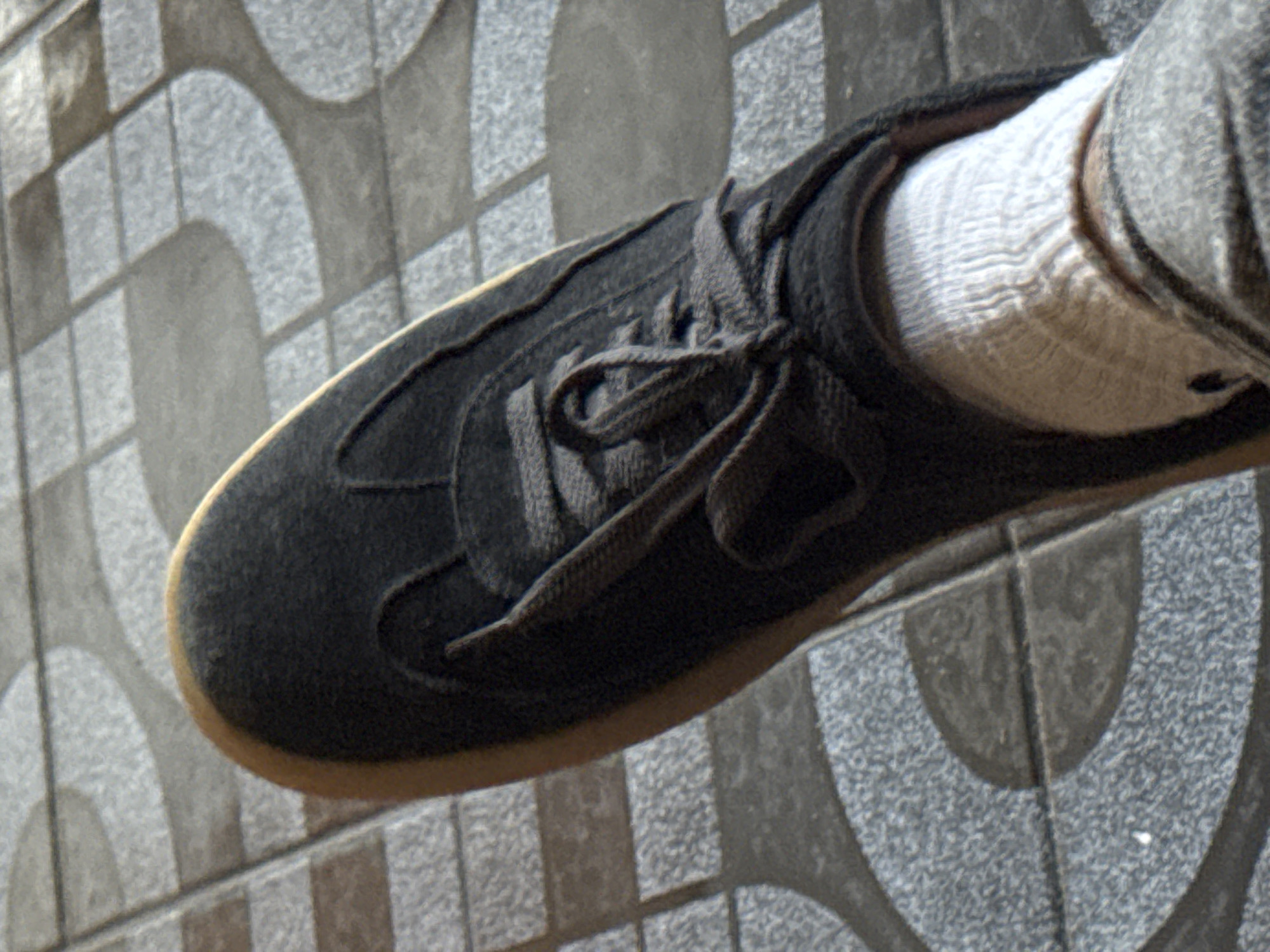 This is a close-up, overhead shot of a person's foot wearing a dark grey, low-top sneaker. The sneaker has laces that are tied in a knot. The sole of the sneaker appears to be a light brown color. The person is wearing a white sock with a subtle ribbed pattern. The background is a tiled surface with a pattern of grey and white semicircles and squares. The lighting suggests it is daytime, possibly outdoors, as there are shadows cast on the tiled surface. There is no visible text in the image. The scene is static, with the focus solely on the shoe and sock.