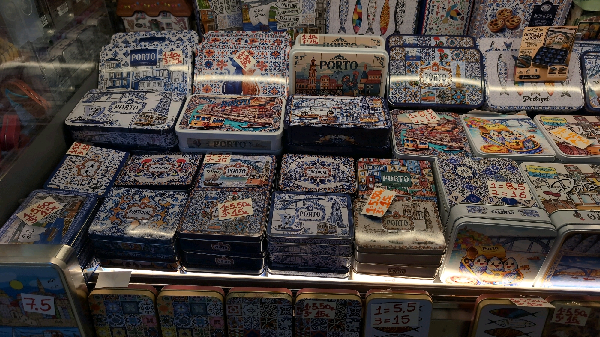 The image displays a retail display of decorative tins, predominantly featuring imagery related to Porto, Portugal. The tins are stacked in multiple rows, showcasing various designs. Many tins are blue and white, with patterns resembling traditional Portuguese tiles (azulejos). Other tins depict landmarks and scenes from Porto, such as trams, bridges, and buildings. Some tins have price tags attached, indicating various prices in euros. One tin in the upper right corner is labeled "INSIDE THE BOX CHOCOLATE CANDIES" and "Sweet Balila," suggesting the tins contain confectionery. The overall environment appears to be a shop or market stall. No people are visible in the image.