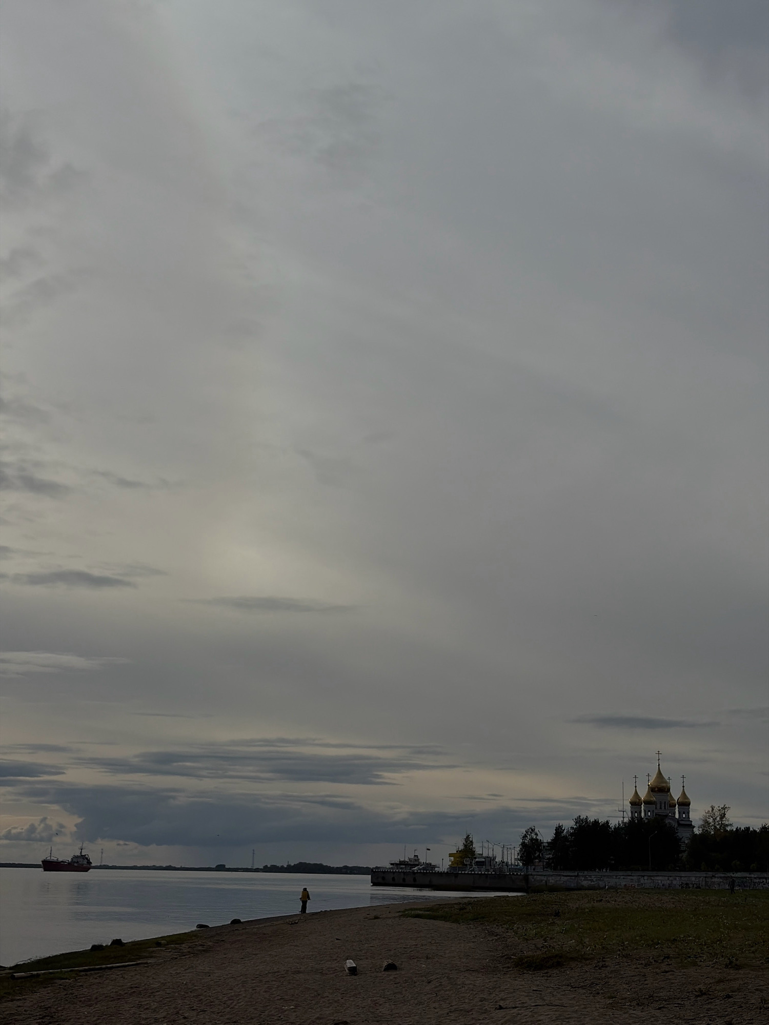 The image depicts a cloudy sky over a body of water, likely the Northern Dvina River in Arkhangelsk, Russia. In the distance, a red cargo ship is visible on the left, and on the right, a large, ornate church with golden domes stands prominently against the treeline. The foreground consists of a sandy embankment with sparse vegetation. A lone figure is seen walking on the embankment near the water's edge. The overall atmosphere is somber, with muted colors and diffused light, suggesting overcast weather.
