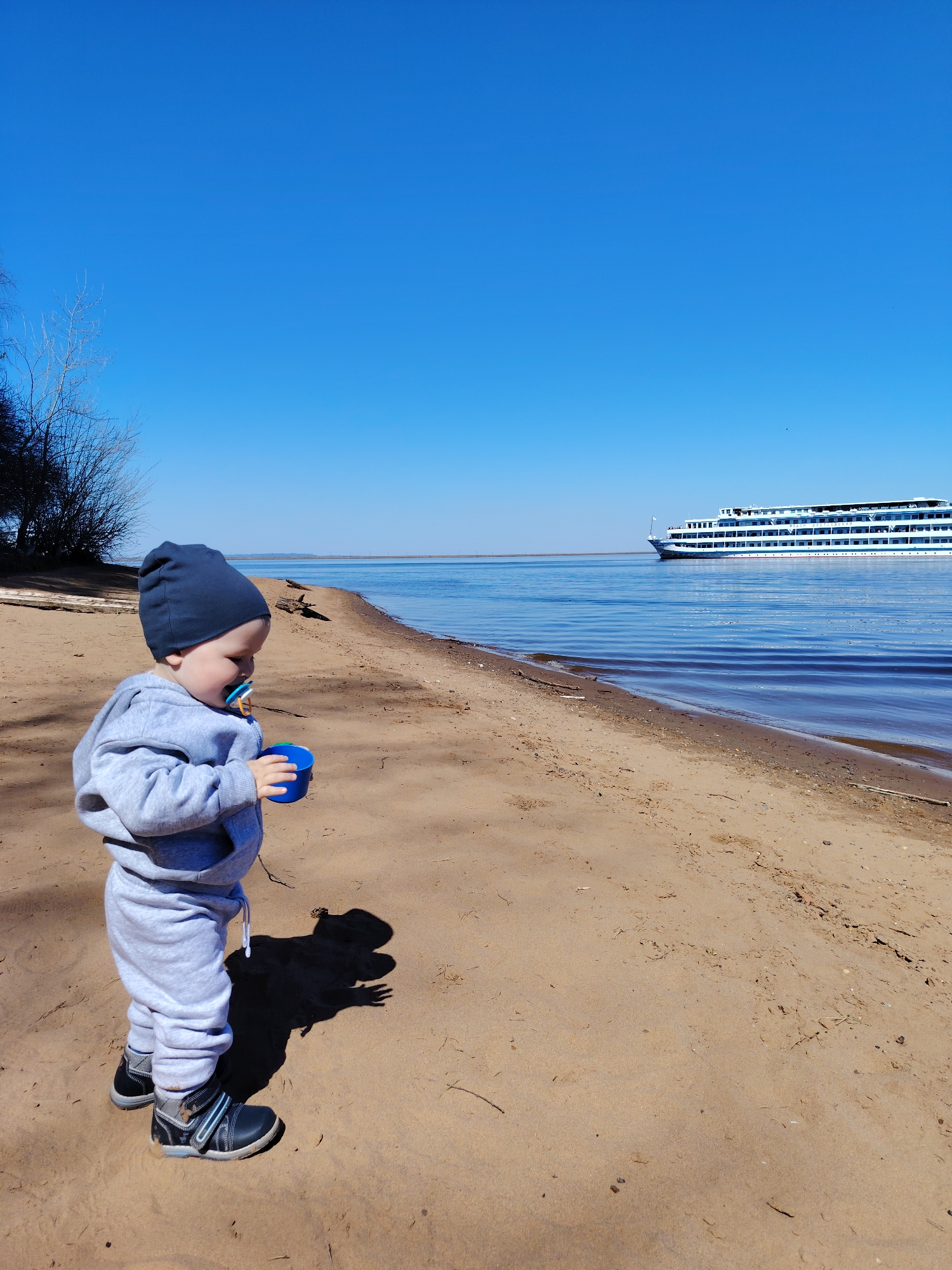 A young child wearing a gray tracksuit, a dark blue beanie, and black shoes stands on a sandy beach, holding a blue cup. The child has a pacifier in their mouth. The beach is located along a body of water, and a large, multi-story cruise ship is visible in the distance on the water. The sky is clear and blue. Bare trees are visible at the left edge of the frame. The child casts a shadow on the sand. The location is the beach in the city of Chaikovsky, Russia.