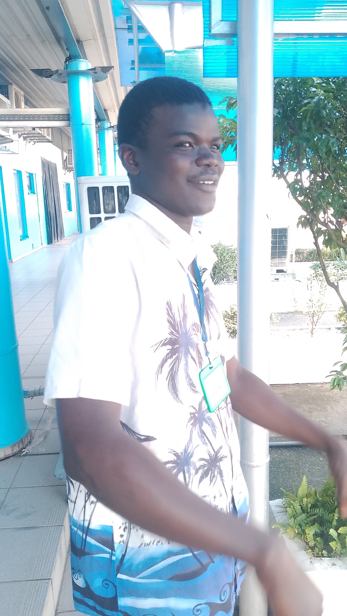 A young Black male is standing outdoors, facing right and slightly away from the camera. He has short dark hair and is smiling. He is wearing a short-sleeved white collared shirt with a palm tree pattern and blue and white patterned shorts. A blue lanyard with a green badge is hanging around his neck. The background features bright blue awnings, light-colored walls, and a large metallic pole. To the right, there is a tree with green leaves and a patch of ferns. The ground appears to be a tiled walkway. The location is likely Orerokpe, Nigeria, given the architectural style and vegetation.