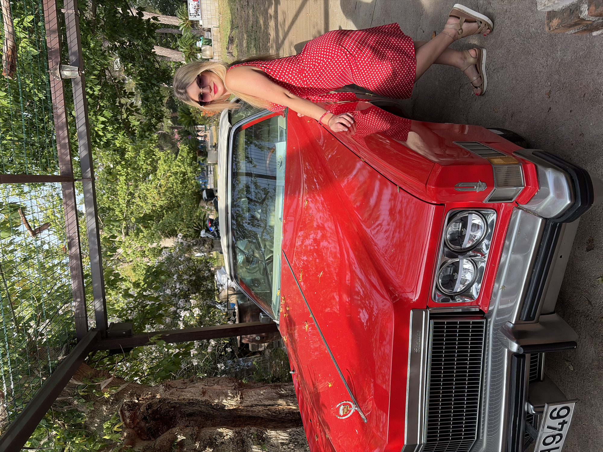 The image shows a woman leaning against a red vintage Cadillac parked outdoors. The woman is blonde, wearing sunglasses, and a red dress with white polka dots. Her right arm rests on the car, and she is wearing sandals. The Cadillac has a white roof and a chrome grille. The license plate number is 1946. The scene appears to be outdoors with trees visible in the background. The image appears to be taken in the city of Budva, Montenegro.