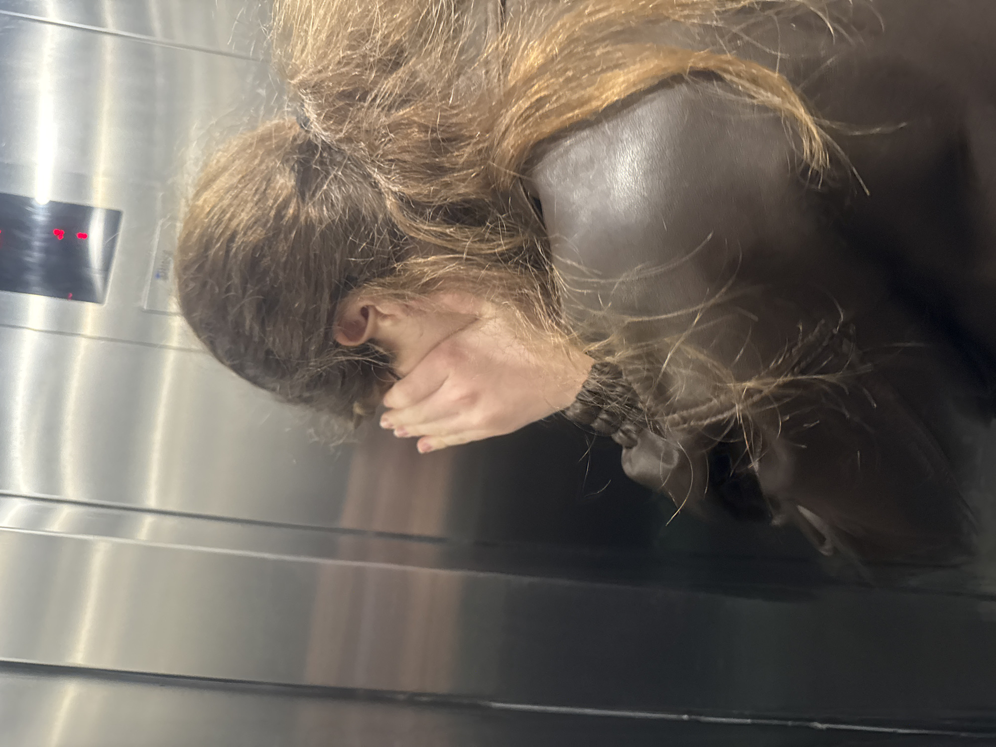 A person with long, brown hair, wearing a dark, possibly leather, jacket, is standing in an elevator. The person's face is partially obscured by their hand. The elevator has a brushed metal interior and a digital display panel showing red numbers. This could be any elevator, and there is no specific information in the image to determine the location in Tbilisi, Georgia.