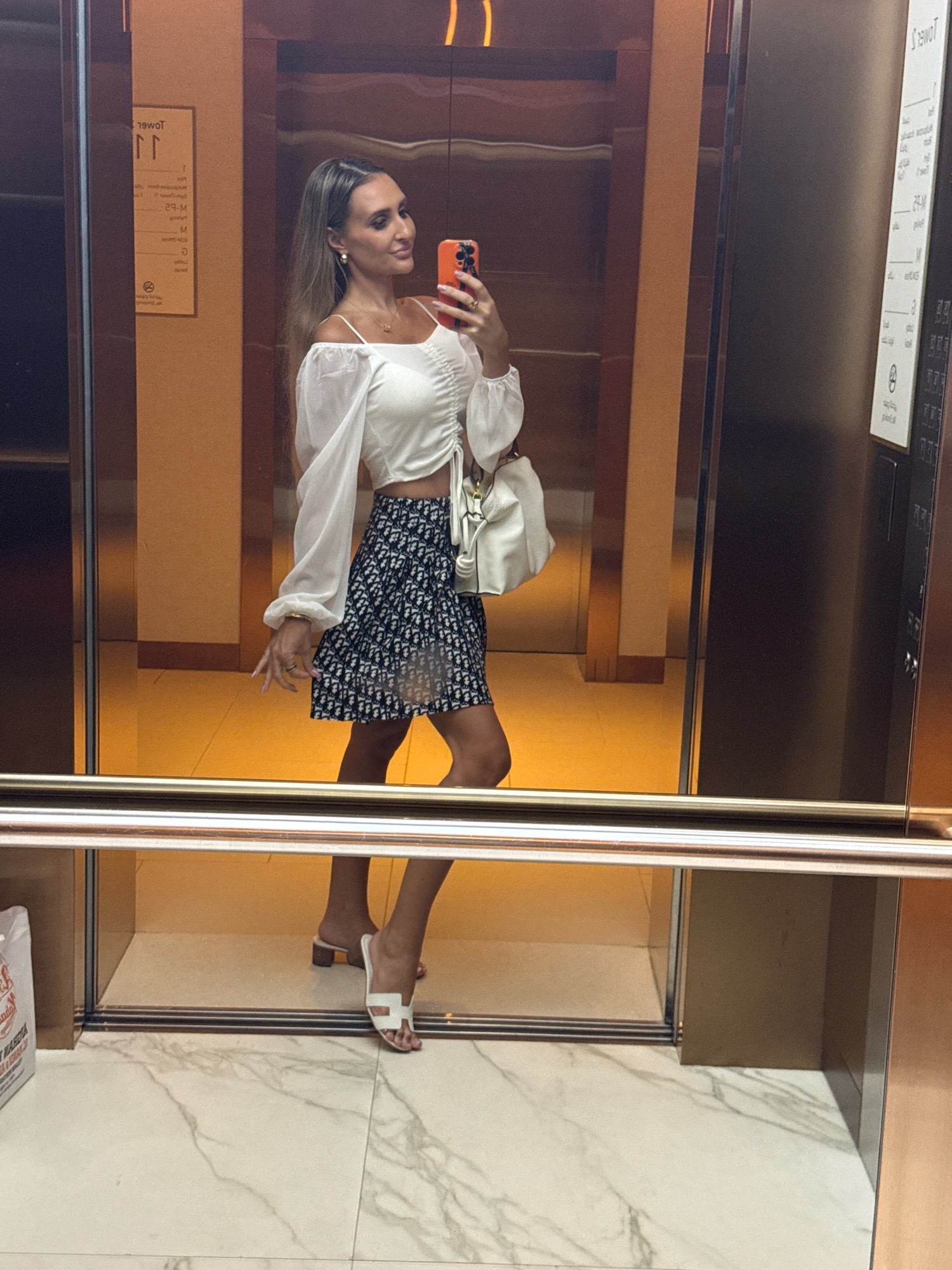 In Dubai, United Arab Emirates, a woman stands in an elevator, taking a selfie with her phone. She is wearing a white cropped top with billowing sleeves and a navy blue and white patterned skirt. She carries a white handbag and wears white sandals. The elevator has mirrored walls and a polished metal railing. The floor is tiled with a white marble pattern. A sign with text in a foreign language is visible on the wall.