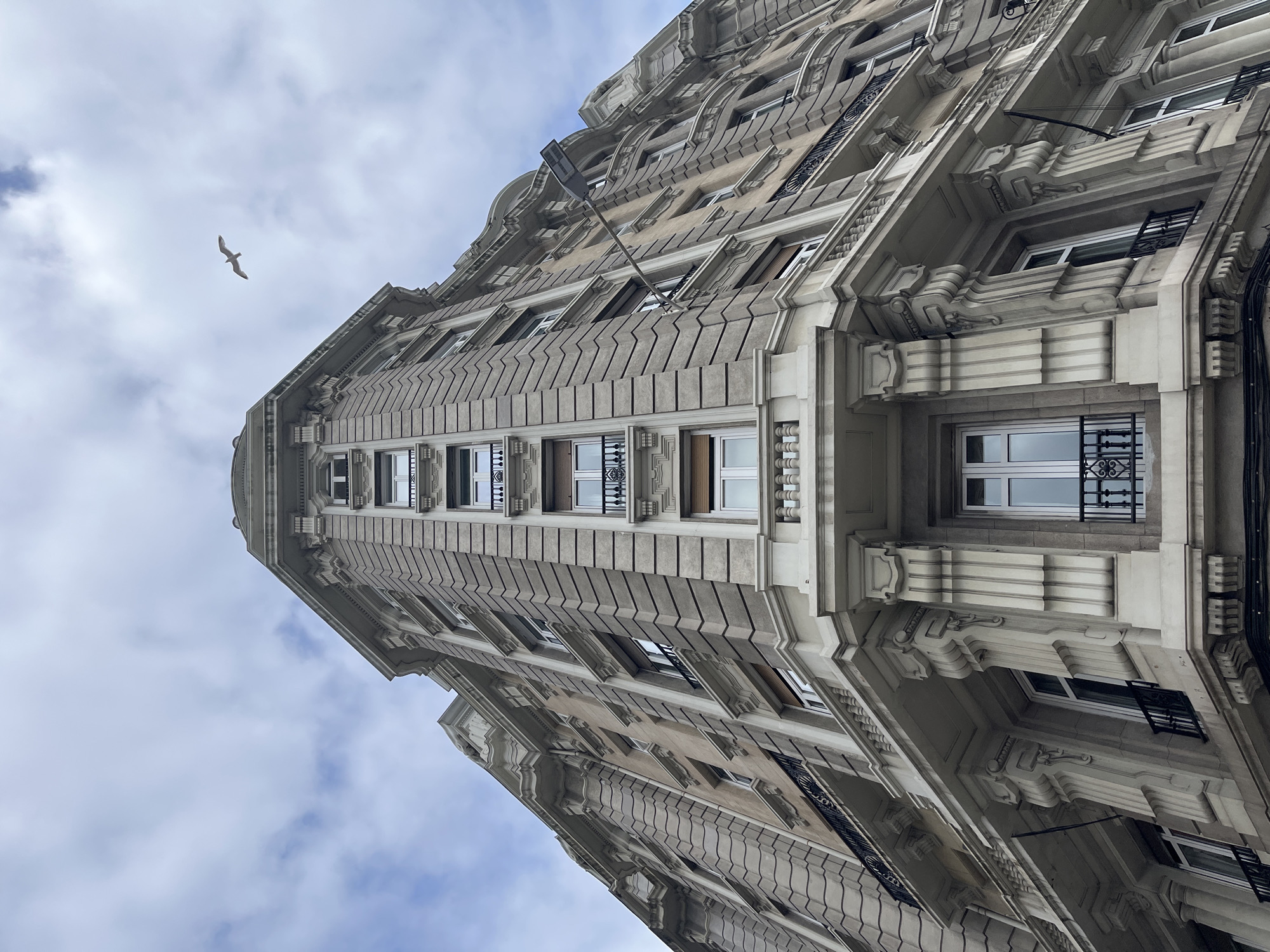 A low-angle shot captures a tall, ornate building in La Coruña, Spain. The building features a light-colored facade with repetitive window patterns and intricate architectural detailing. A bird is visible in the upper left of the frame against a cloudy sky. The building appears to be a historic structure.