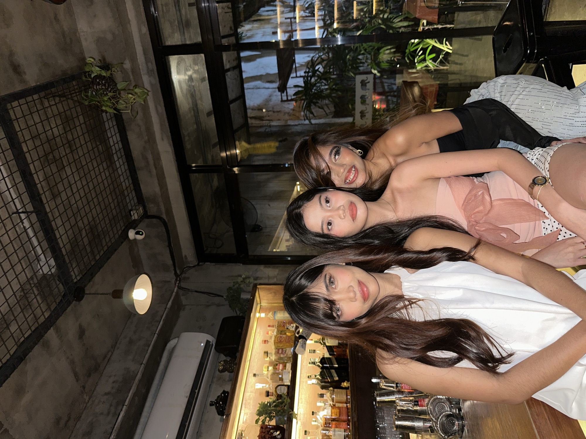 Three young women are posing for a photo in what appears to be a dimly lit bar or lounge. The setting features exposed concrete ceilings, industrial-style metal shelving, and a bar stocked with various bottles and glasses. One woman in the foreground wears a white halter dress, while the woman next to her is in a pink strapless dress with a bow detail. The third woman is in a black off-the-shoulder top and sparkly pants, leaning in from behind. Plants are visible in the background, along with reflections in the glass partition, suggesting an urban environment. The overall ambiance is casual and social, likely a night out in Davao City, Philippines.